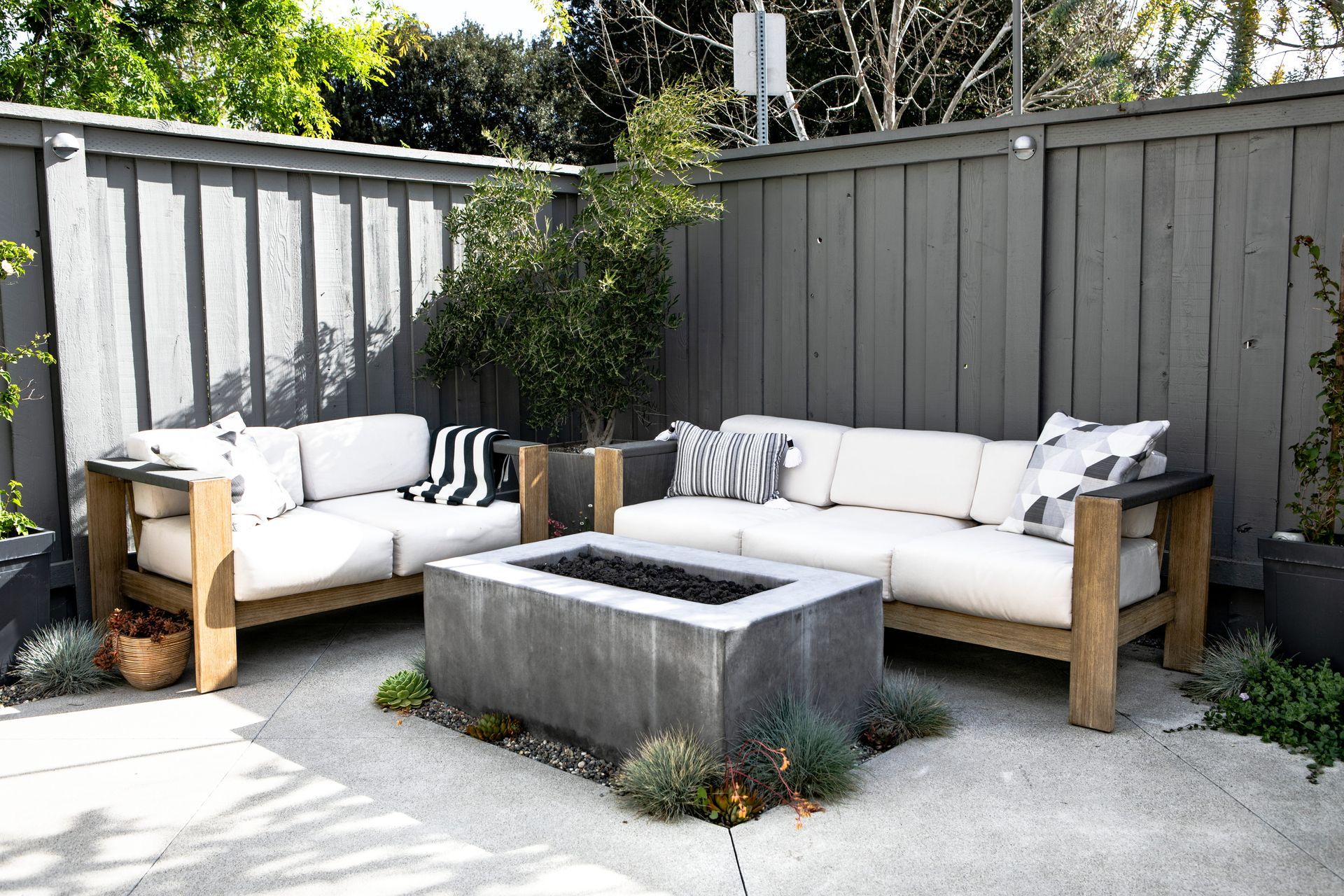 Outdoor patio with two white cushioned sofas around a concrete fire pit, bordered by gray wooden fencing.