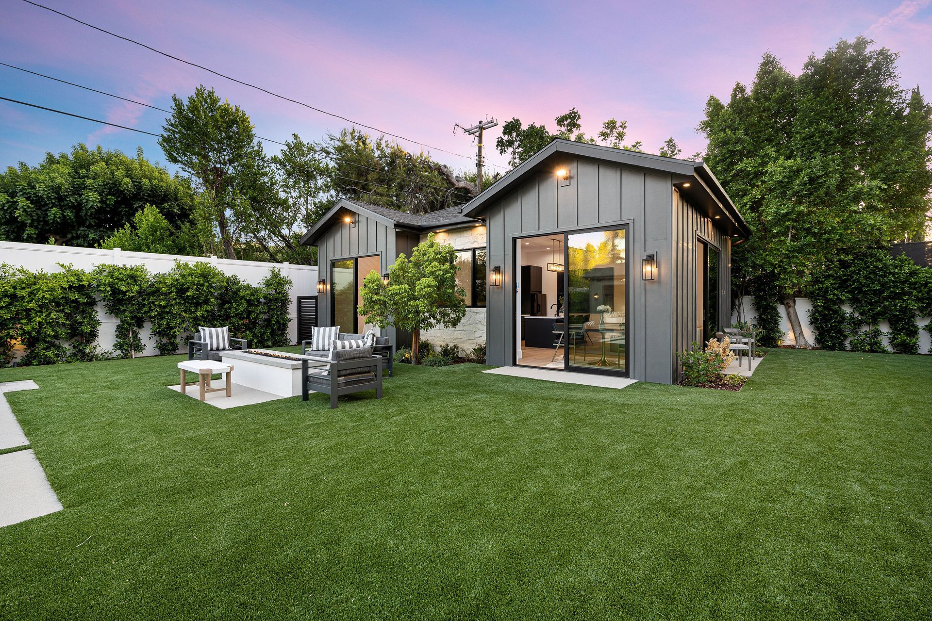 Modern backyard with a small gray cabin, patio seating, and a lush green lawn at sunset