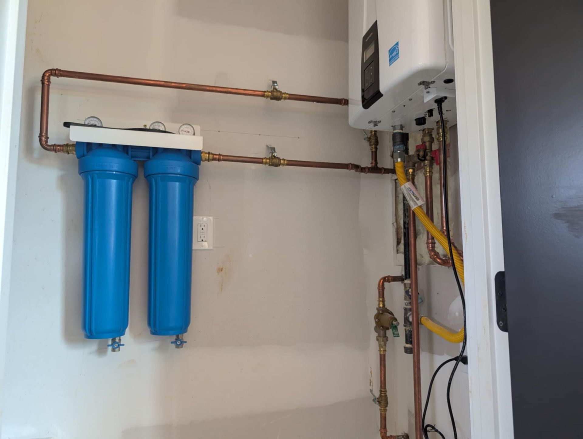 Water filtration system with two blue canisters and a tankless water heater, copper pipes, and gas line.