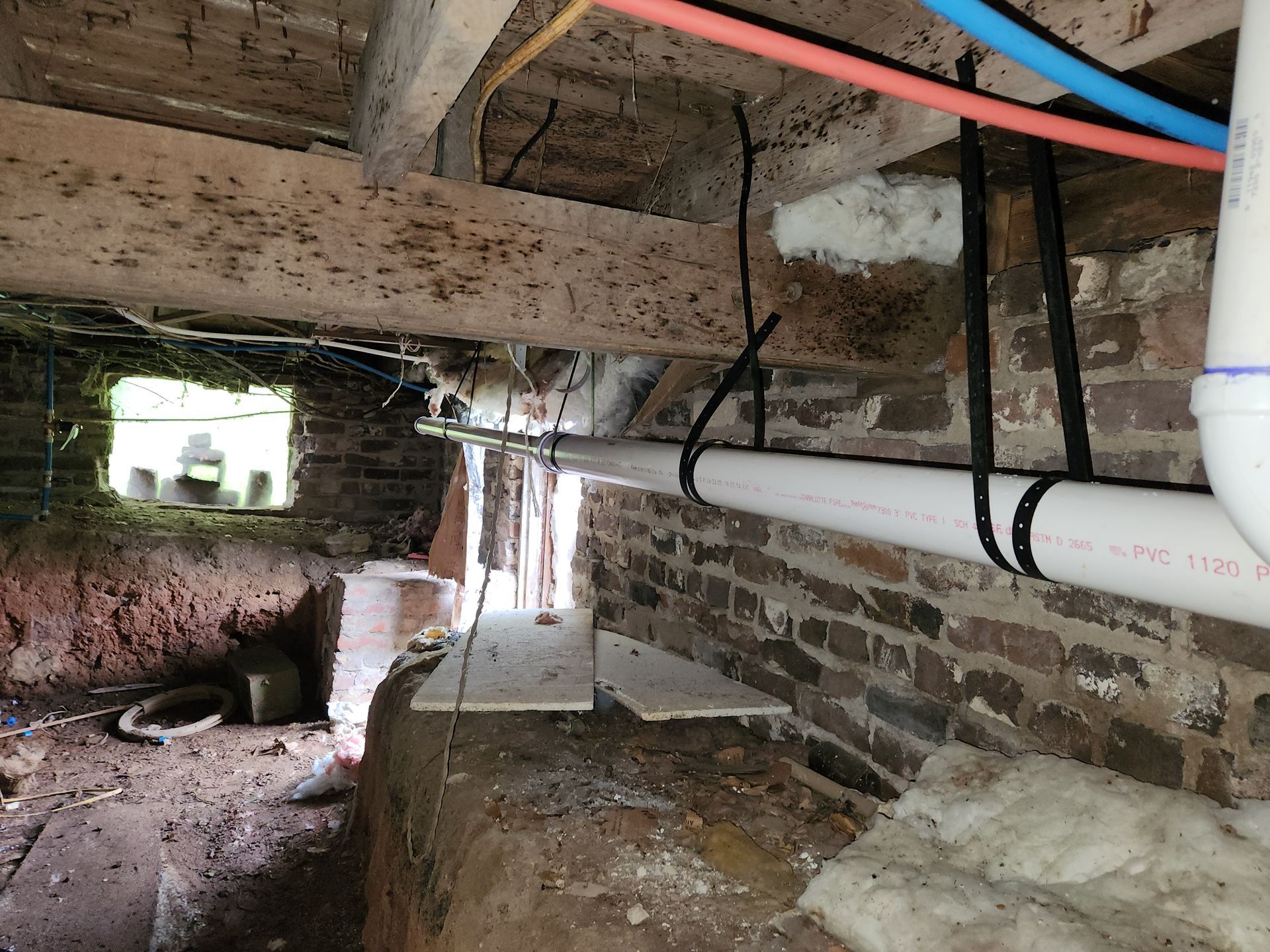 Under-house view showing brick foundation, wooden beams, plumbing pipes, and electrical wiring.
