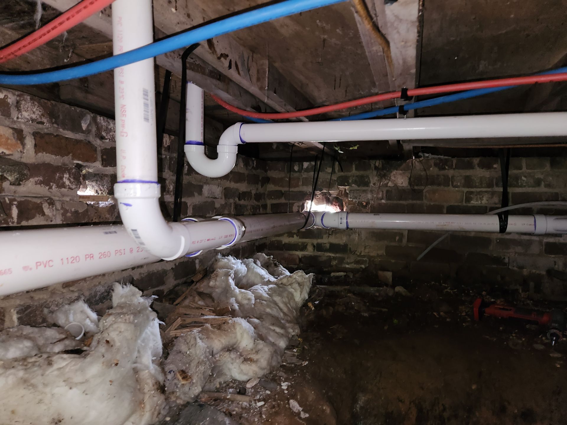 White PVC pipes and red/blue tubes snake across a brick foundation, possibly for plumbing.