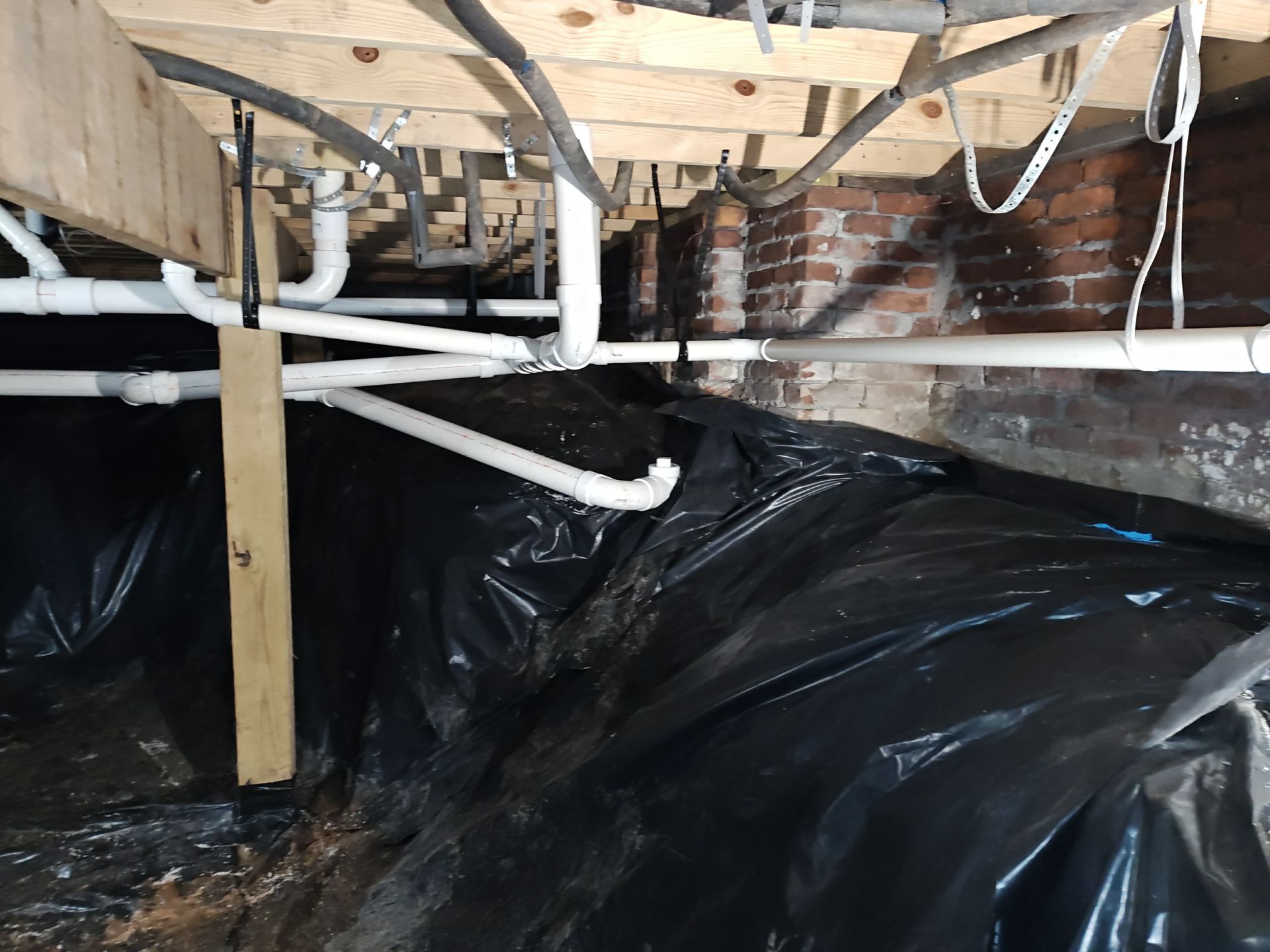 Underneath a building, black plastic sheeting covers the ground, with pipes and brick walls visible above.