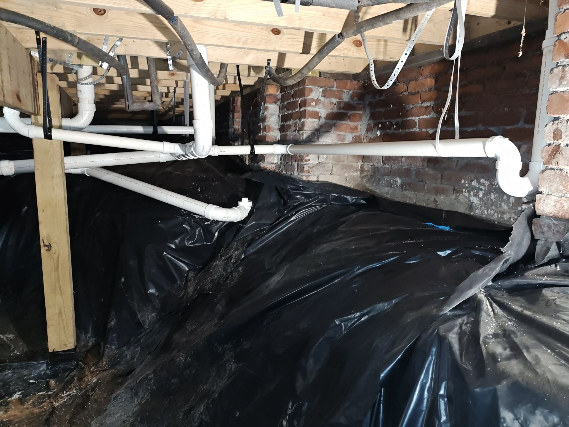 Black plastic sheeting in a crawl space, covering the ground and brick foundation with white pipes overhead.