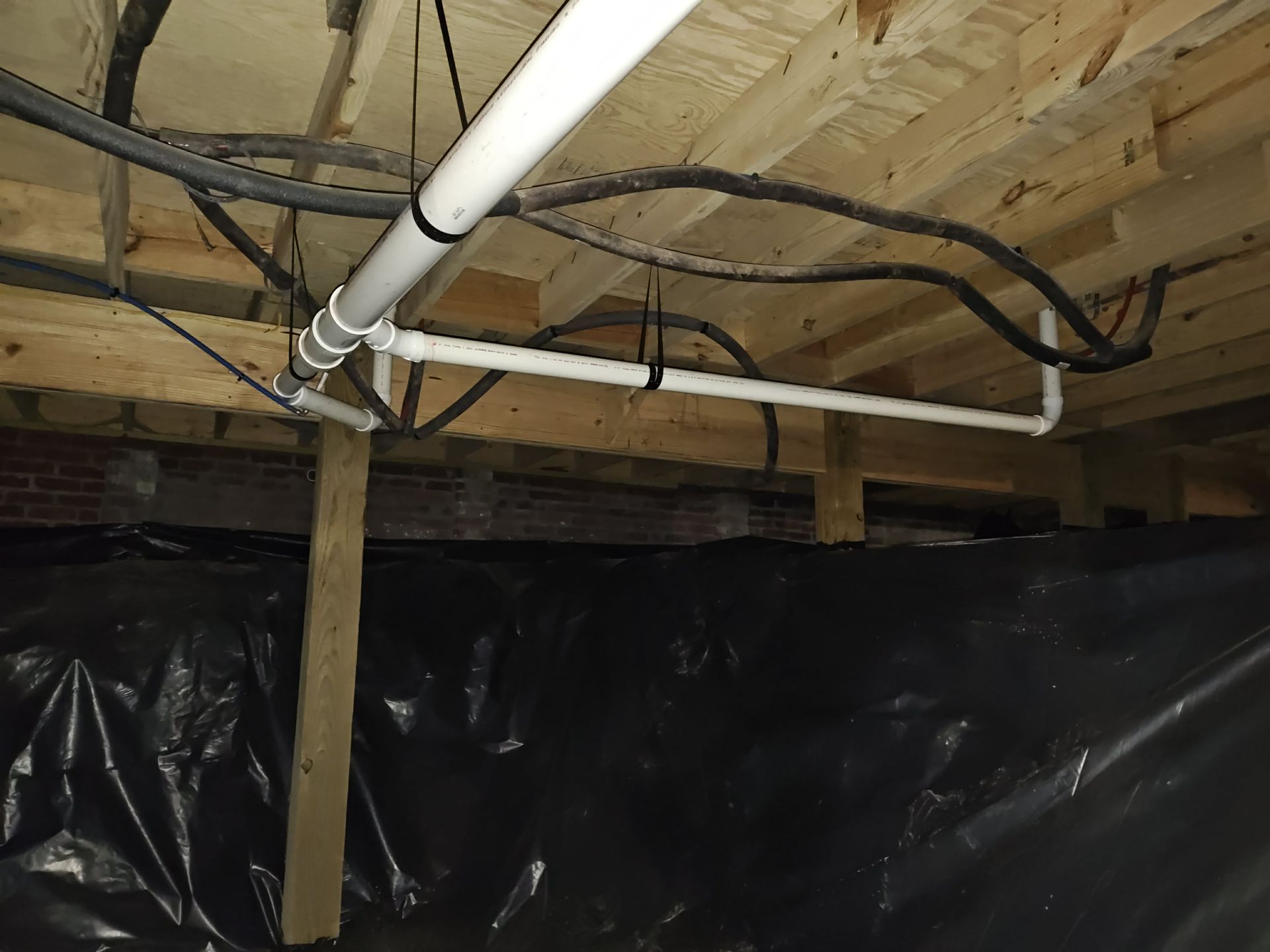 View of a crawlspace with white PVC pipes, electrical wiring, black plastic, and wooden beams.