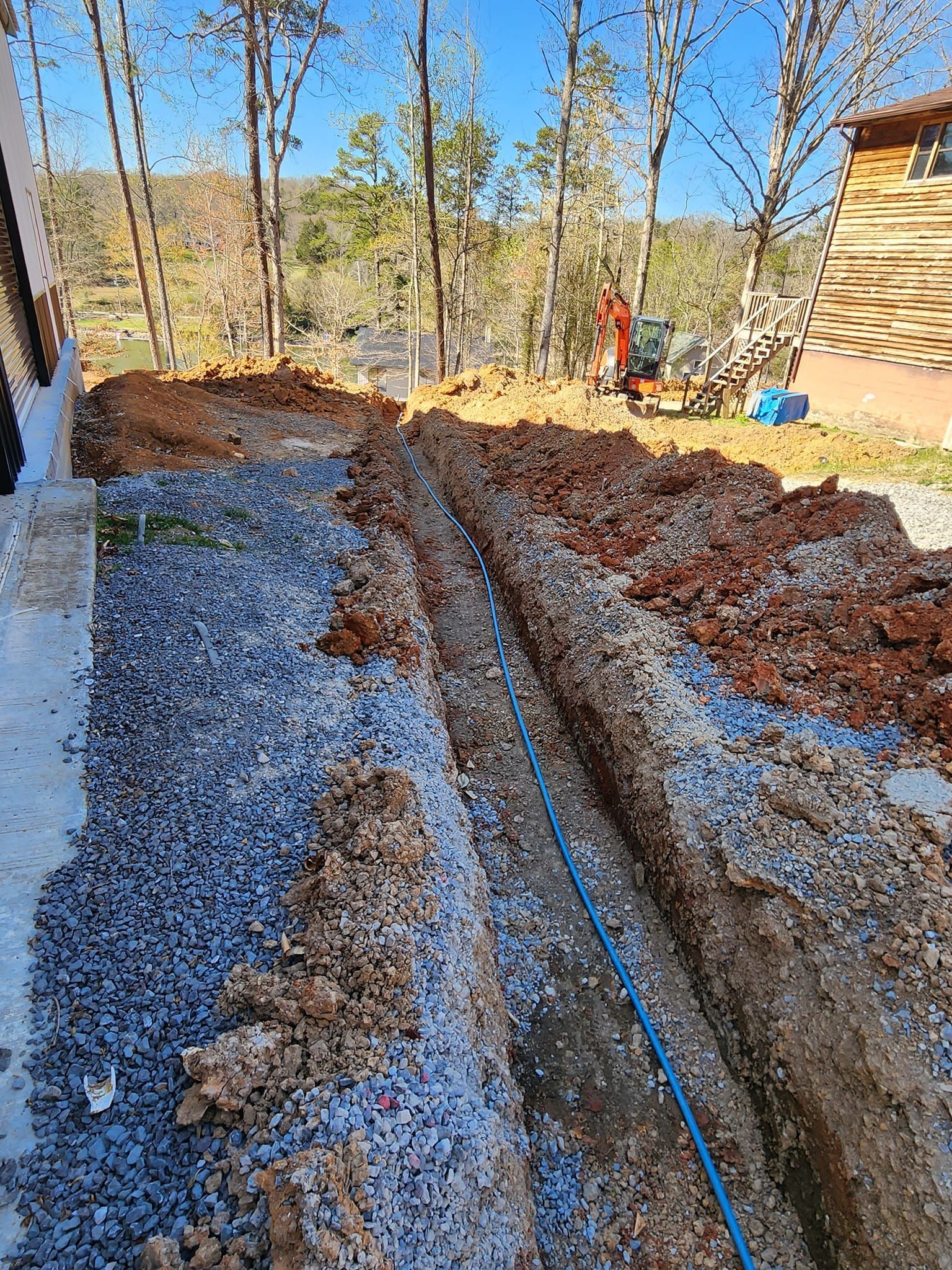 Trench dug for utility lines next to a building; blue pipe visible, surrounded by gravel and soil.