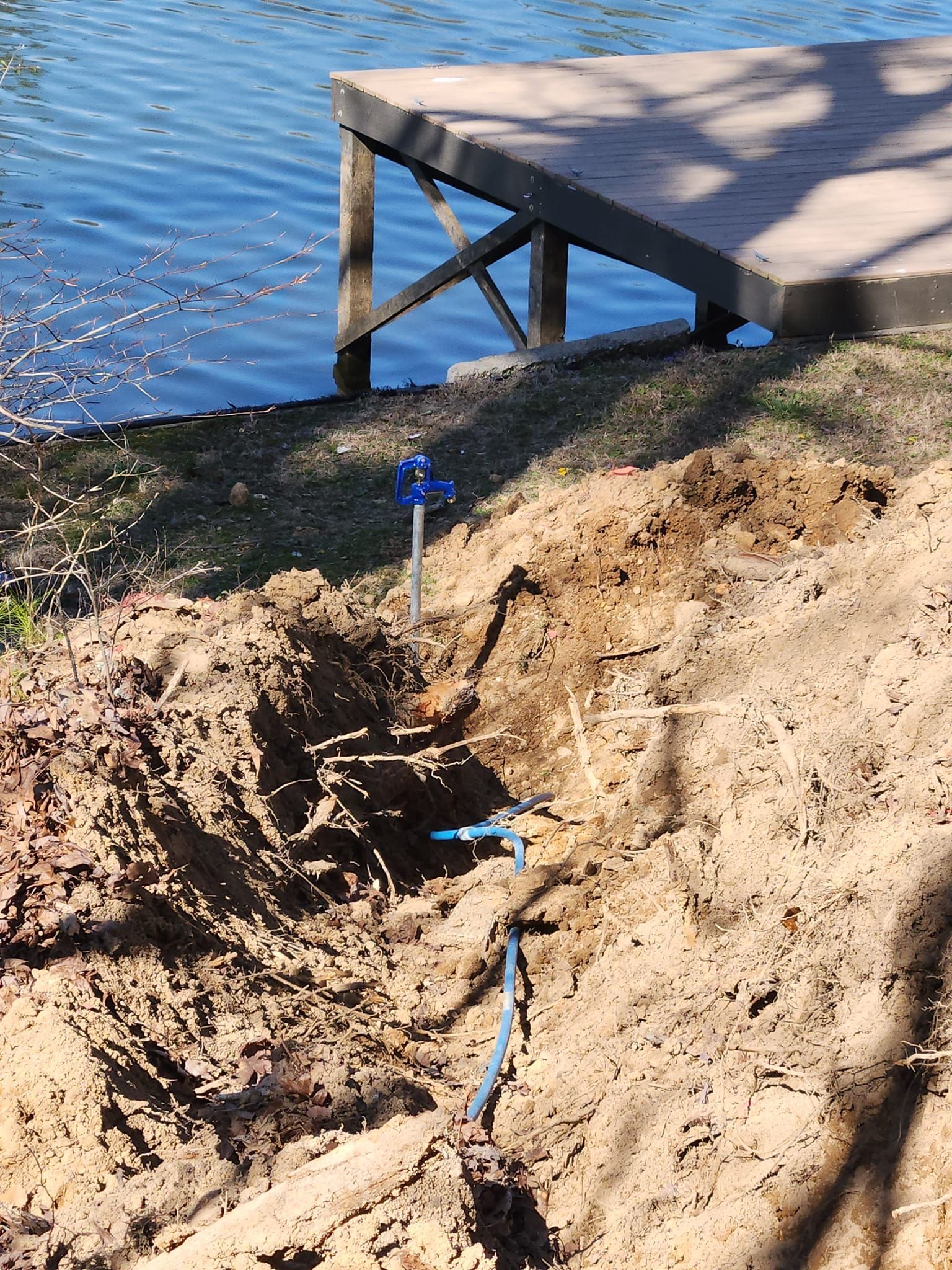 Trench dug near a lake, with blue pipes and a water valve; a wooden dock is visible.