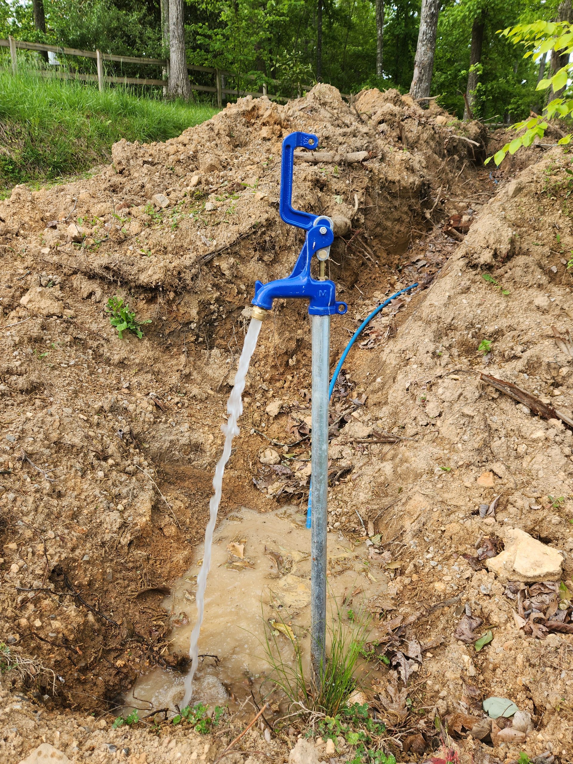 Blue hand water pump spurting water into a trench dug into a dirt mound, in a wooded area.