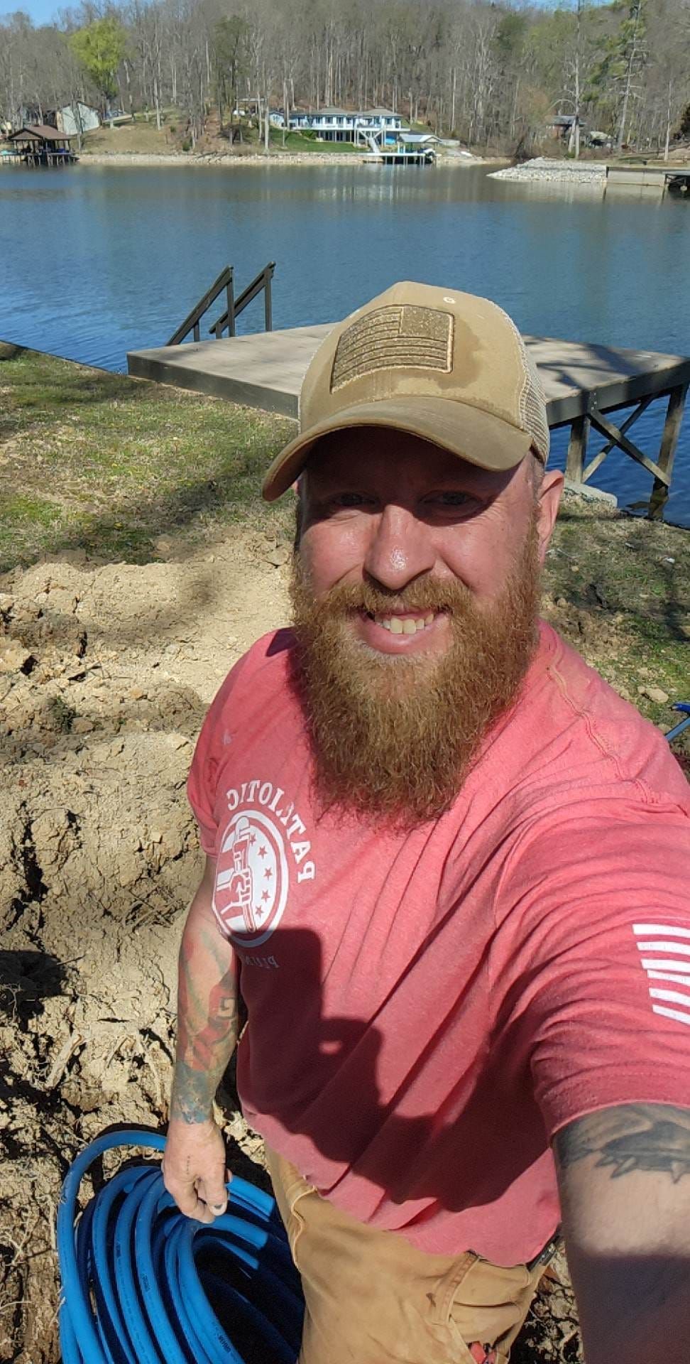 Man with red beard and hat by a lake smiling, holding blue hose.