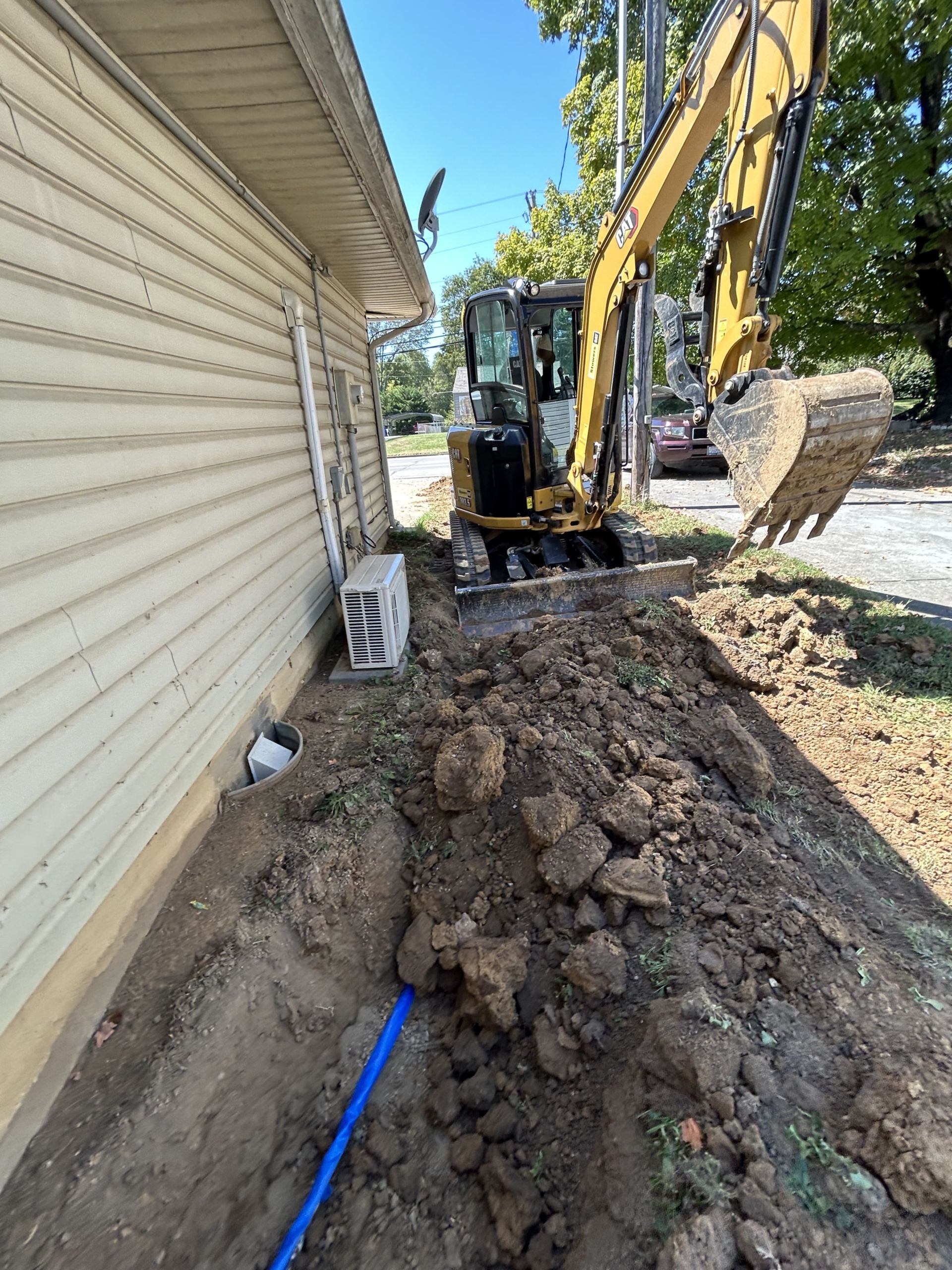 Excavator digging trench beside building; blue pipe visible in trench. Sunny day.