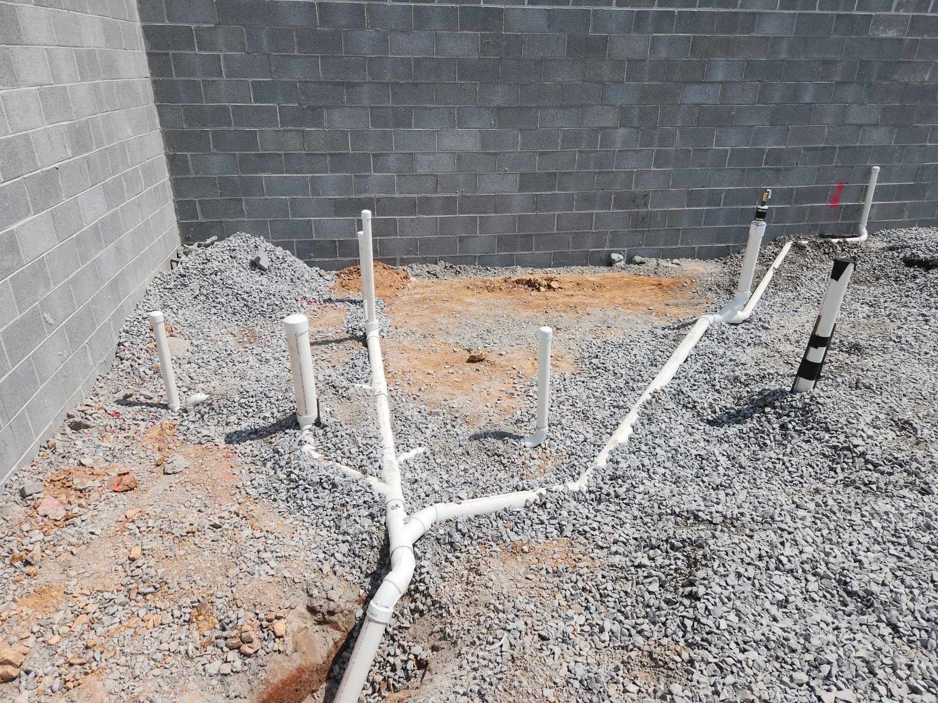 White PVC plumbing pipes on gravel ground, near a cinder block wall, during construction.