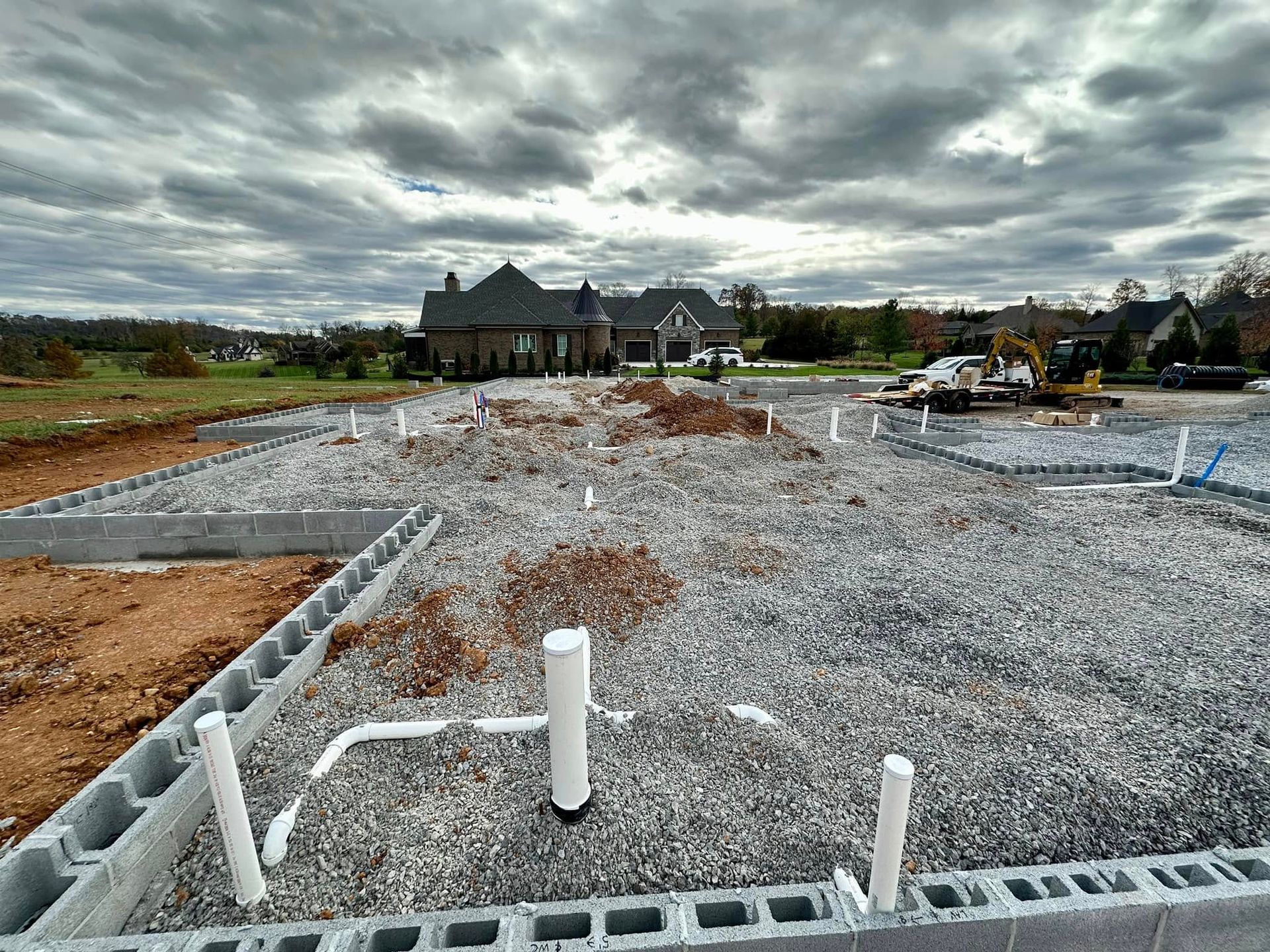 Construction site of a house; cinder block foundation with gravel, plumbing pipes, and cloudy sky.