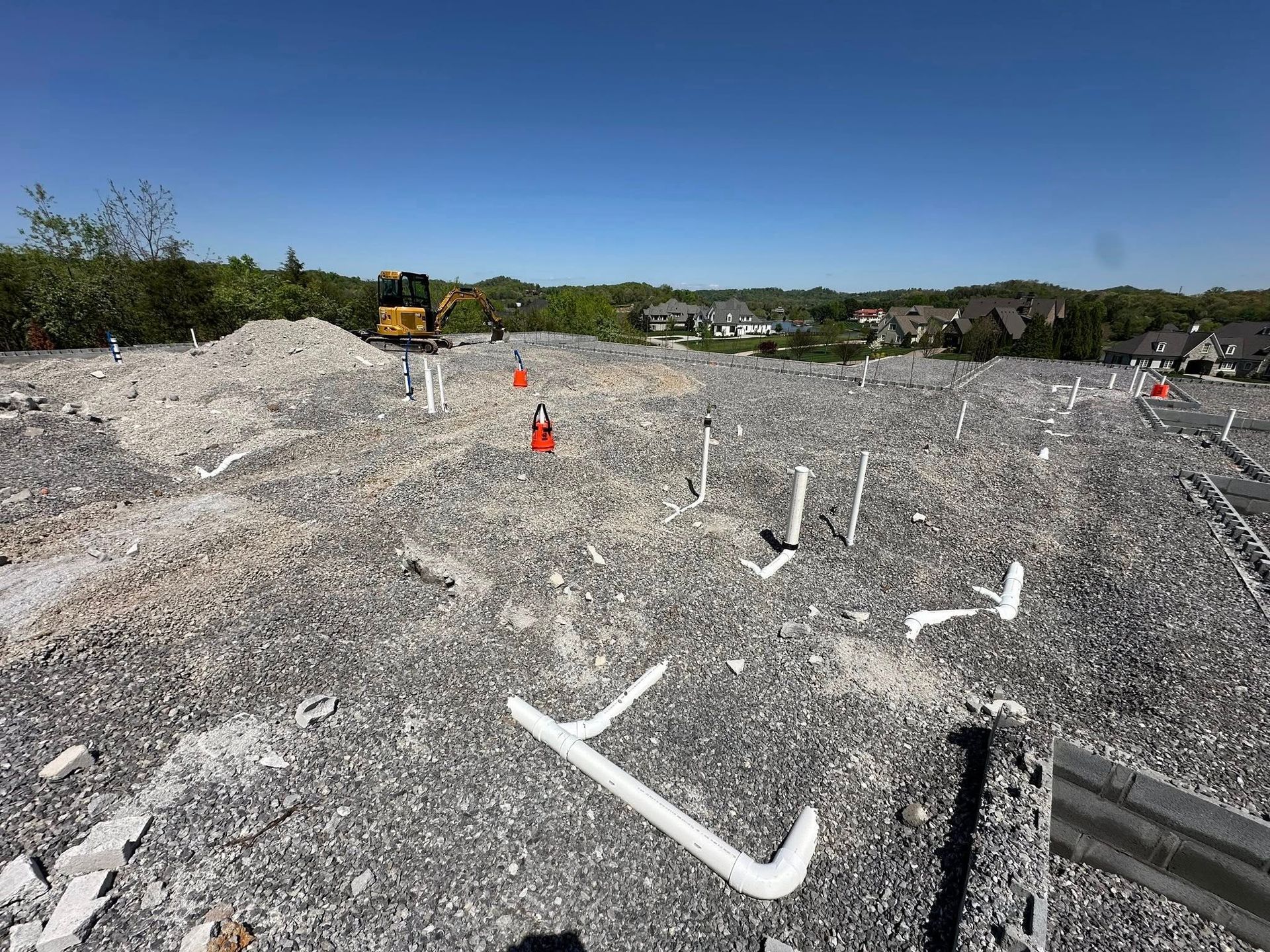 Construction site with gravel, pipes, and an excavator. Clear blue sky, houses in the distance.