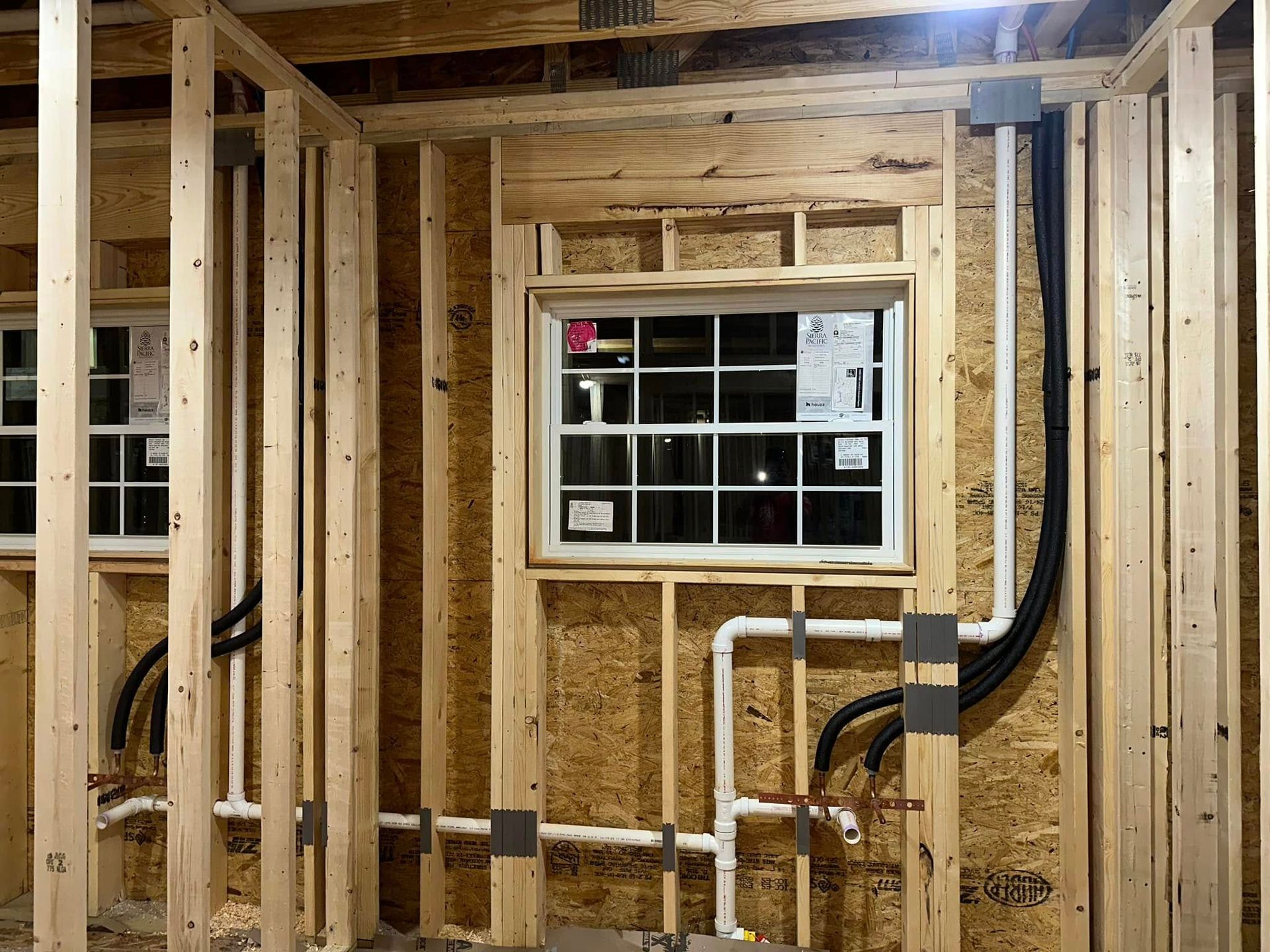 Wooden wall framing with a window and visible plumbing and electrical conduits.