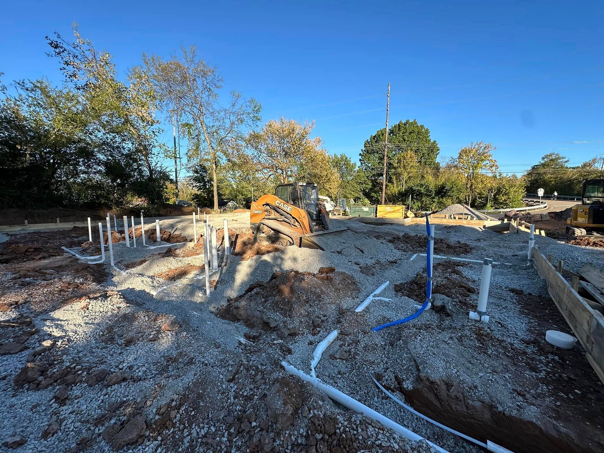 Construction site with plumbing pipes, gravel, and an excavator under a blue sky.