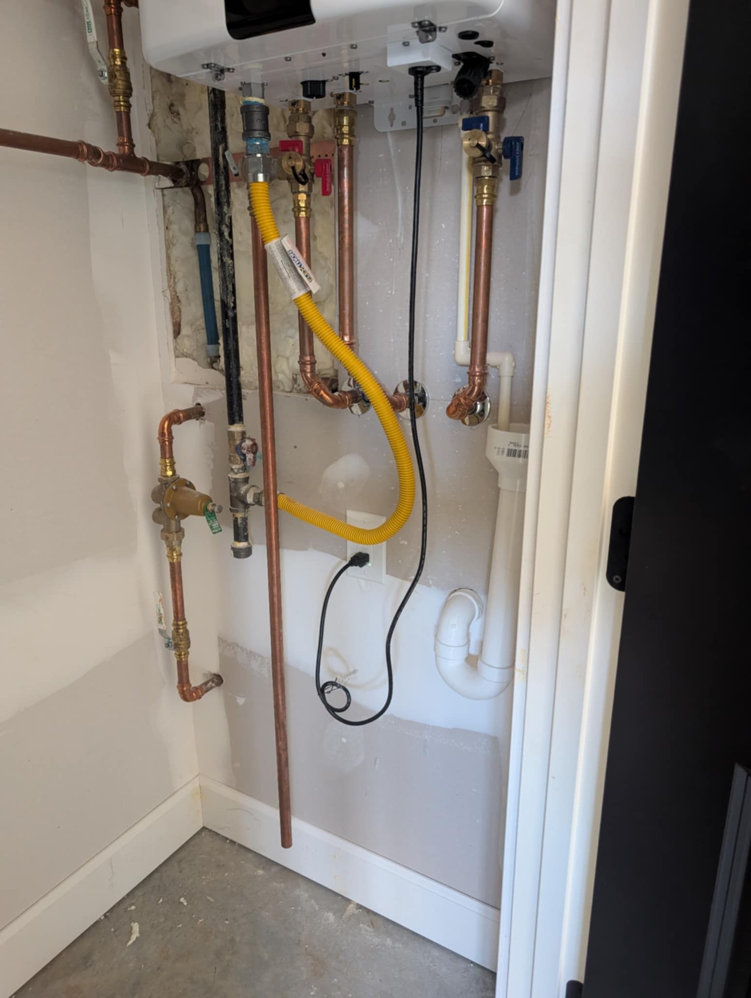 Piping and a water heater in a narrow, white-walled utility closet with copper pipes, a yellow gas line, and a black electrical cord.