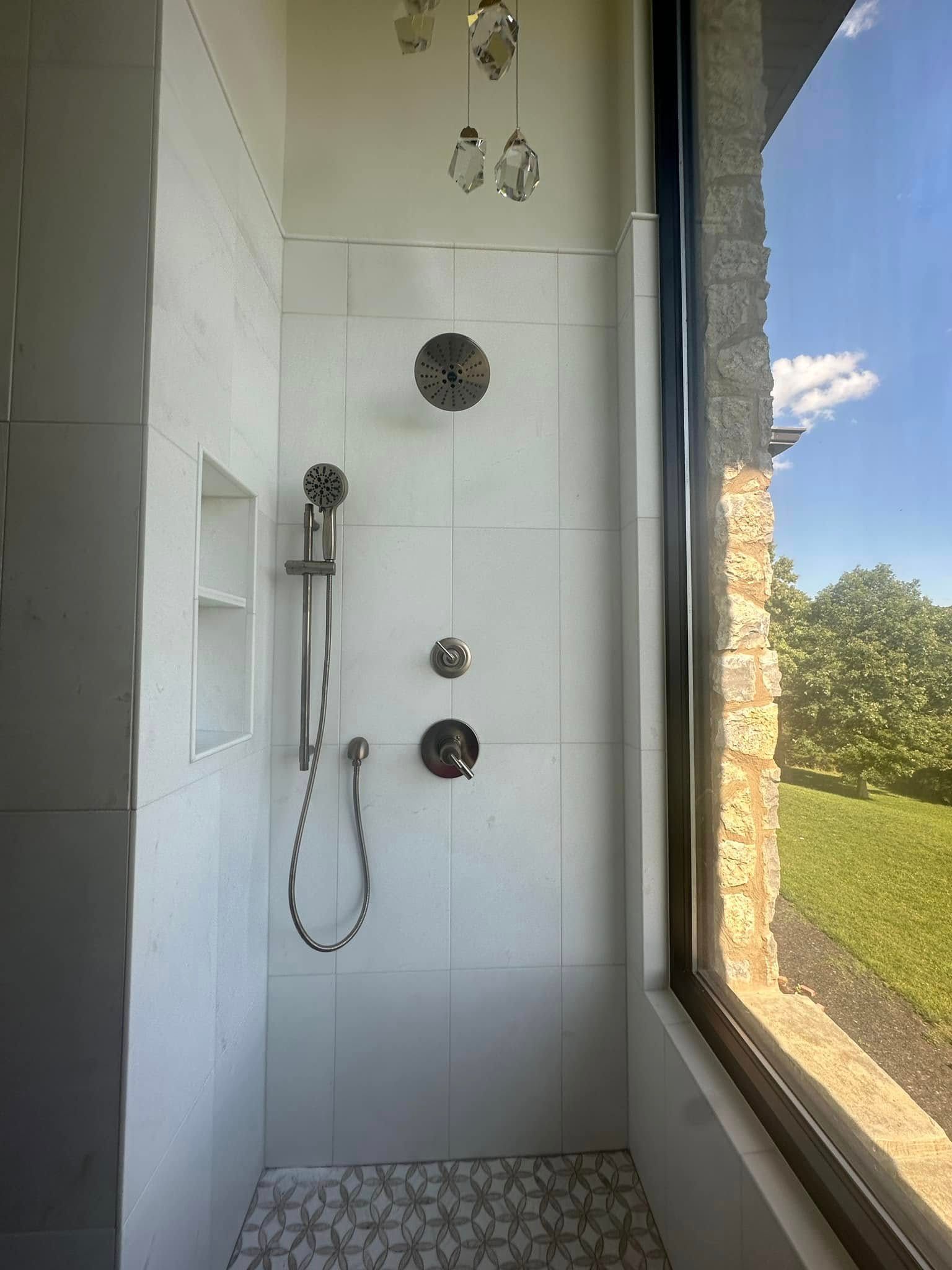 Shower with white tile walls, pebble floor, and large window overlooking greenery.