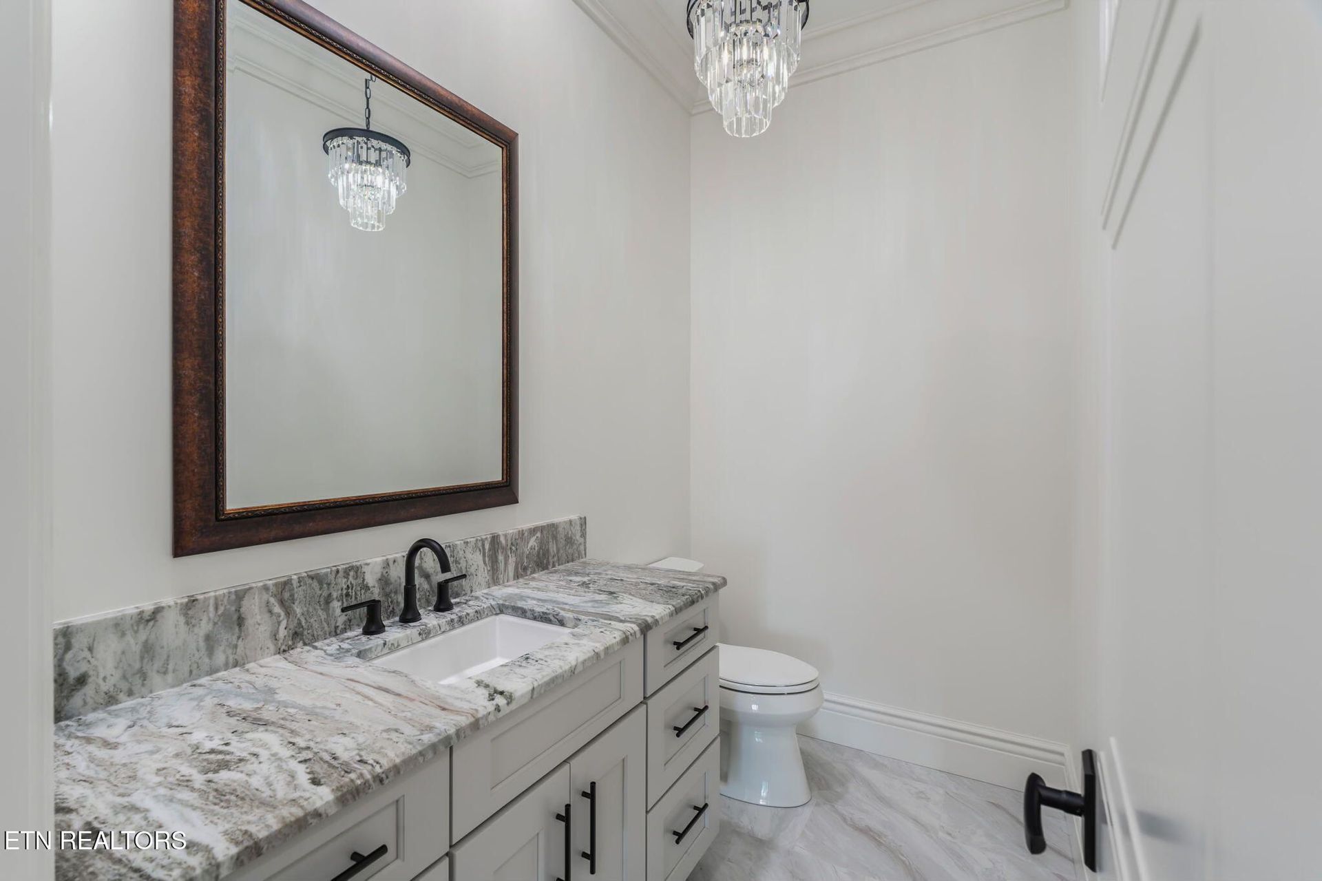 Powder room with white walls, marble countertop, large mirror, and crystal chandelier.