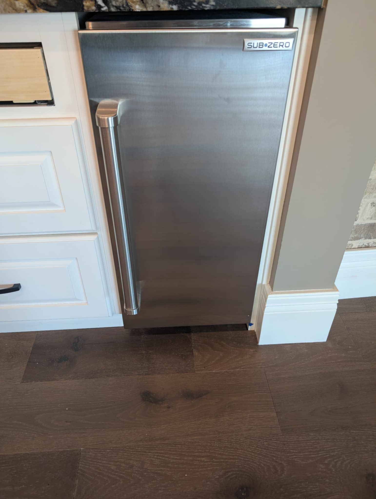 Stainless steel ice maker built into white cabinetry, next to a gray wall and dark wood flooring.