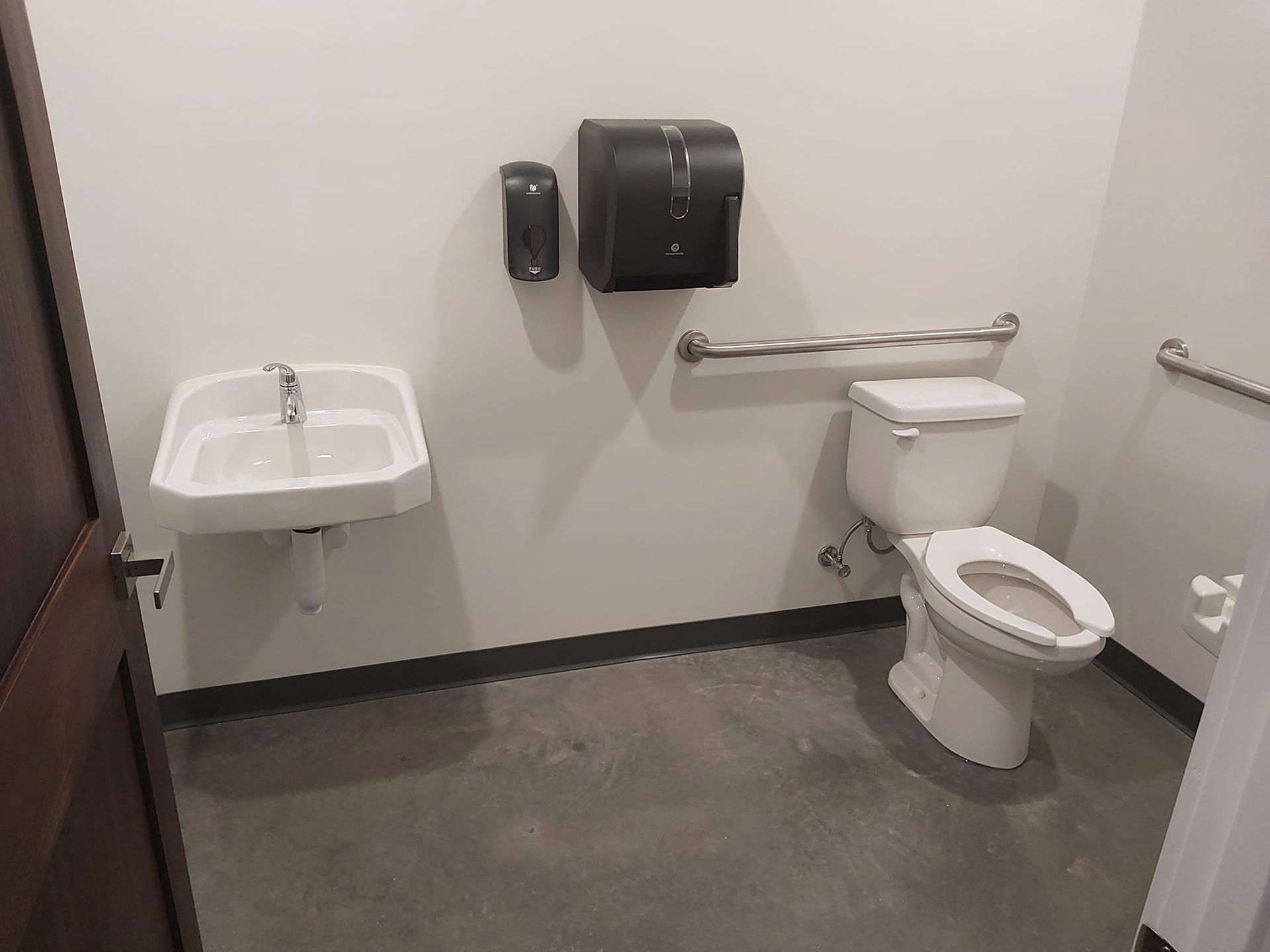 A small, accessible public restroom with a sink, toilet, hand dryer, and grab bars.