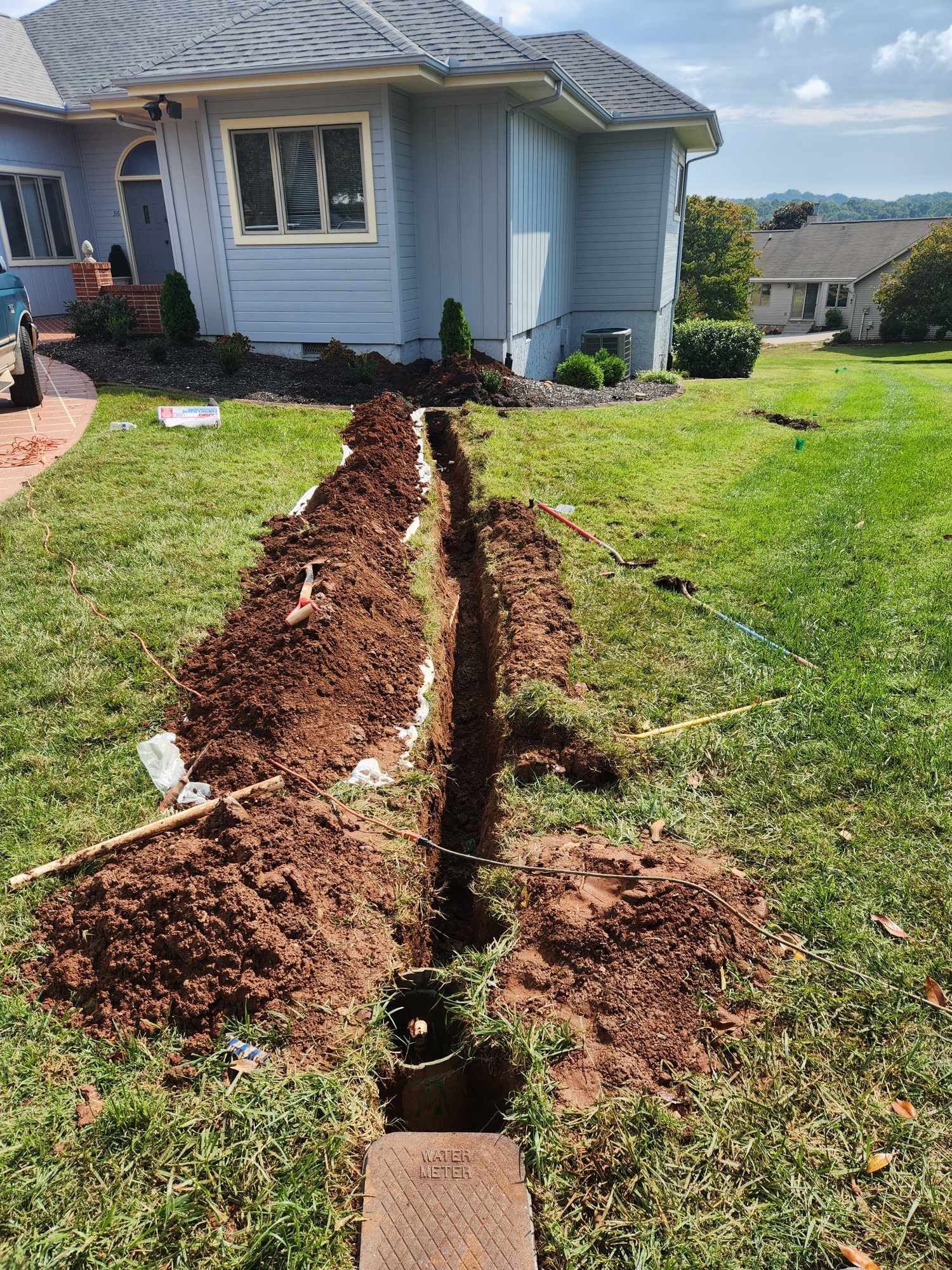 A trench dug in a grassy yard leading to a building with light blue siding.