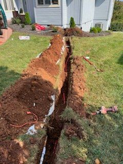 Trench dug in a lawn near a light blue house. Exposed orange and white wires and copper pipes are visible in the trench.