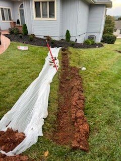 A trench dug in a lawn near a house, covered with white tarp and a red shovel.