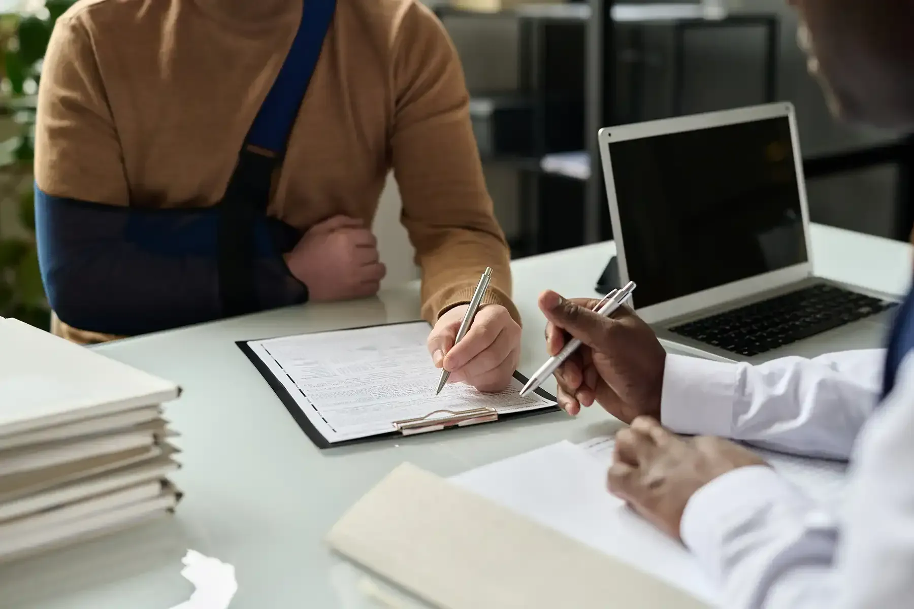 Person with arm in sling signing paperwork at a desk with a doctor, laptop, and file folders.