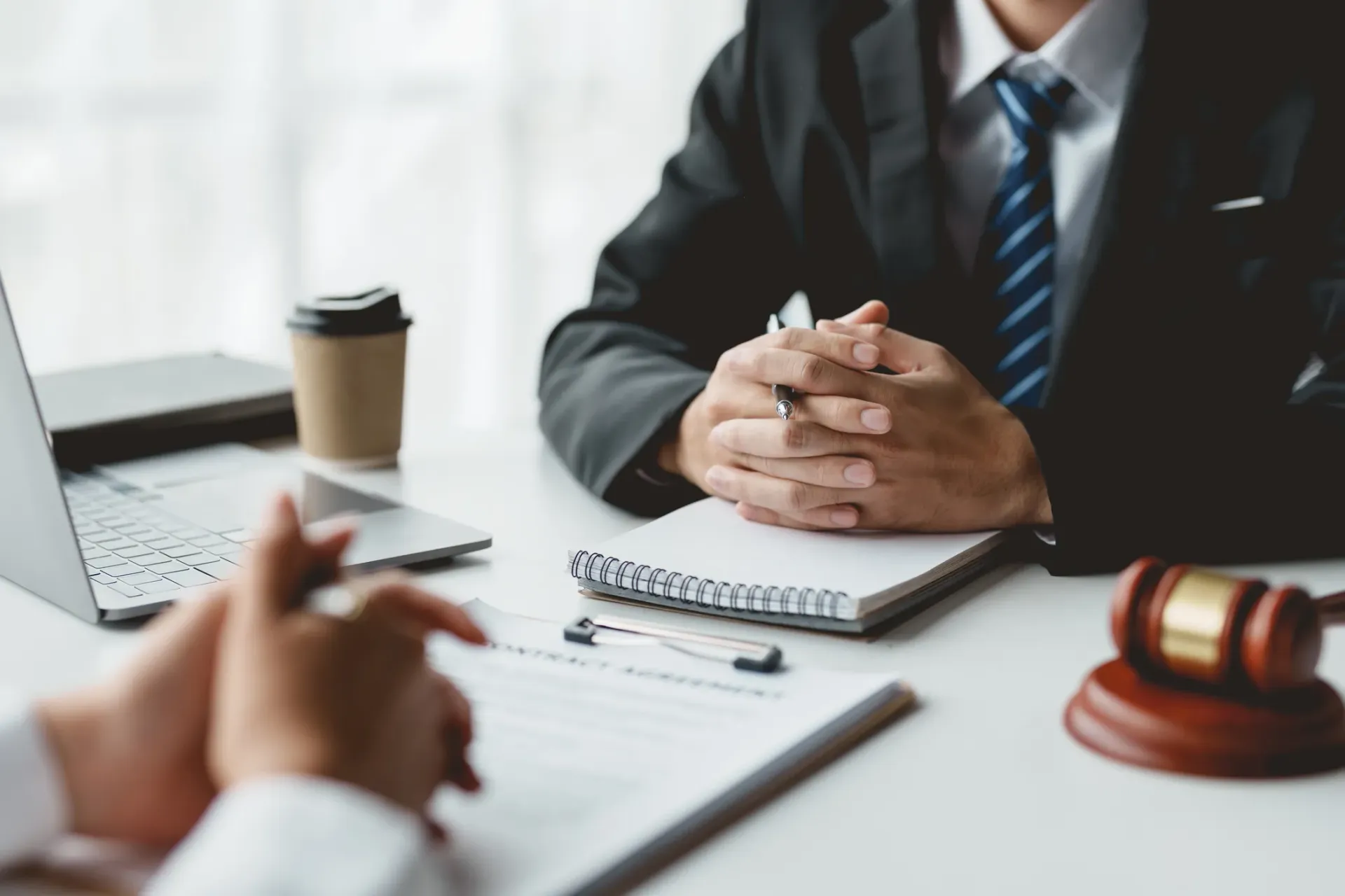 Two people in business attire at a table with a laptop, documents, notepad, gavel, and coffee.