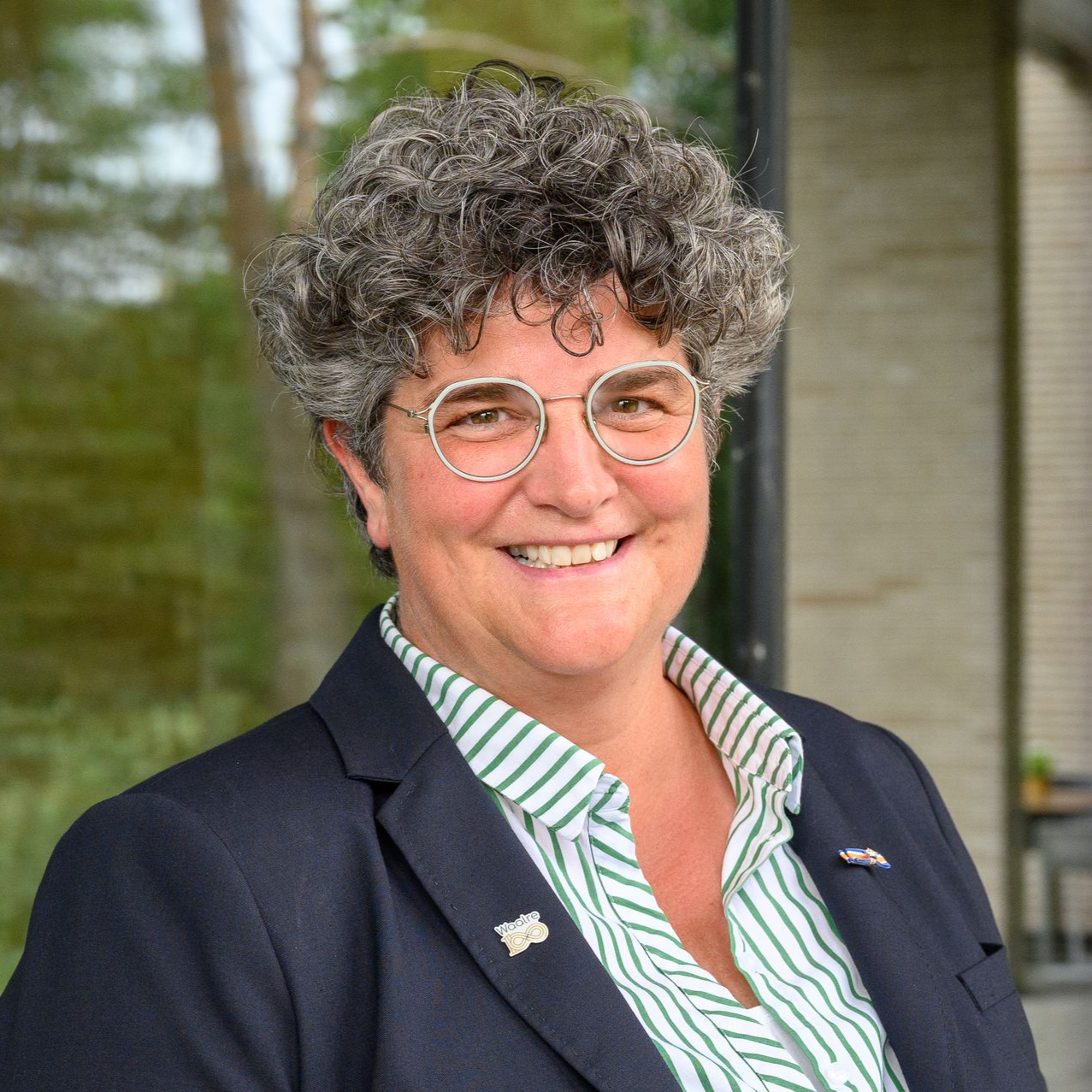 Een vrouw met krullend grijs haar, een bril, een gestreept shirt en een blazer lacht buiten.