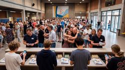 Mensen onder elkaar op een receptie, sommigen omhelzen elkaar, terwijl ze drankjes en kleine schaaltjes met eten vasthouden.