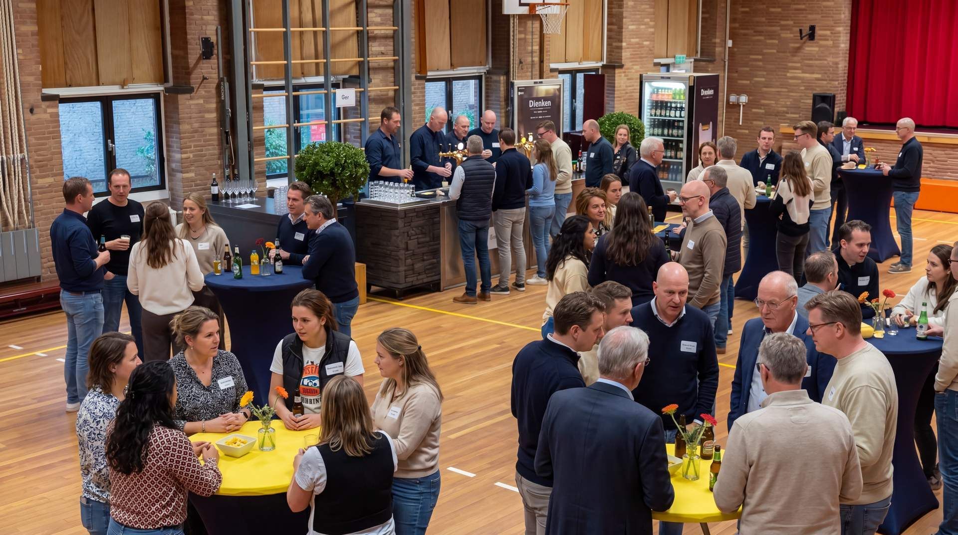 Mensen mengen zich onder elkaar rondom gele tafelkleden tijdens een levendig netwerkevenement in een bakstenen zaal.
