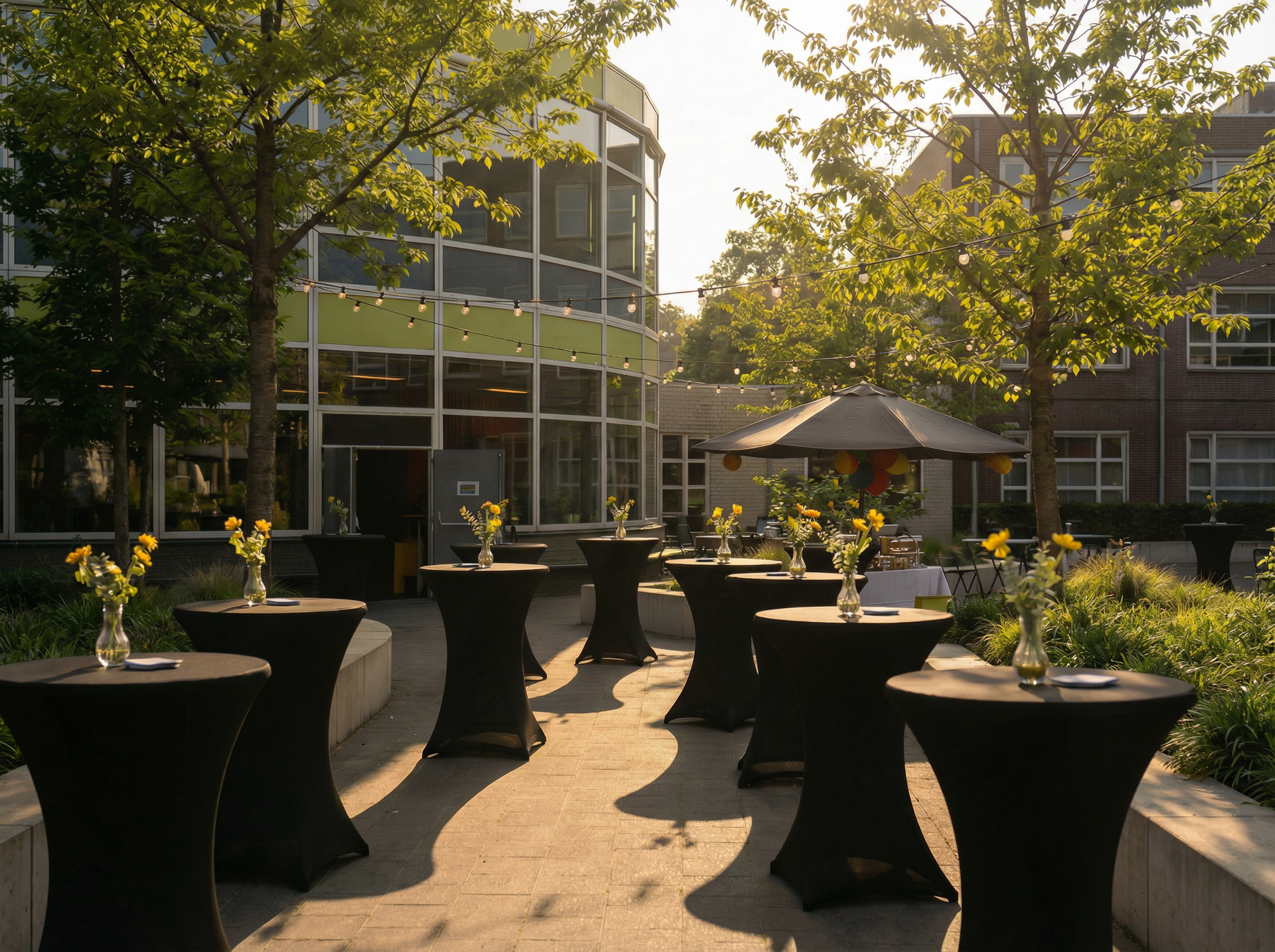 Buitenevenement met zwarte cocktailtafels en gele bloemen, vlakbij een modern gebouw met bomen.