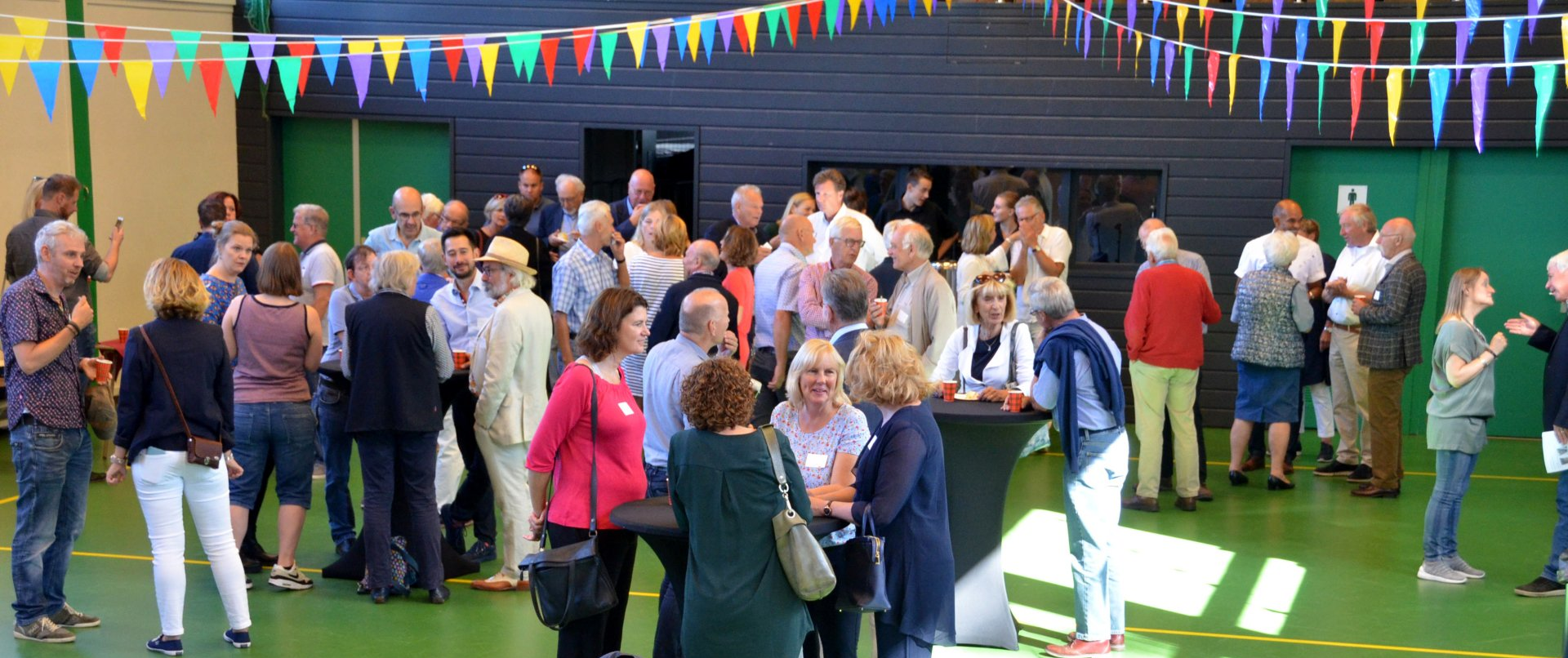 Een grote groep mensen die gezellig samen zijn in een felverlichte binnenruimte, met kleurrijke spandoeken boven hun hoofden.