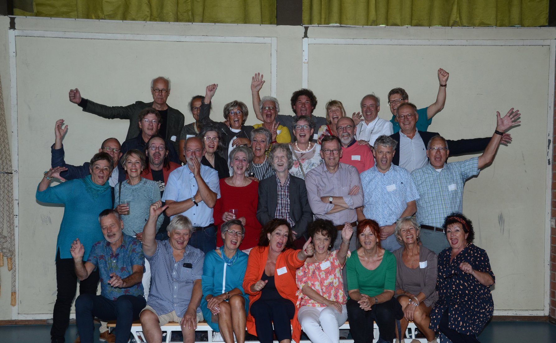 Een groep mensen zwaait en lacht. Ze staan en zitten voor een beige muur.