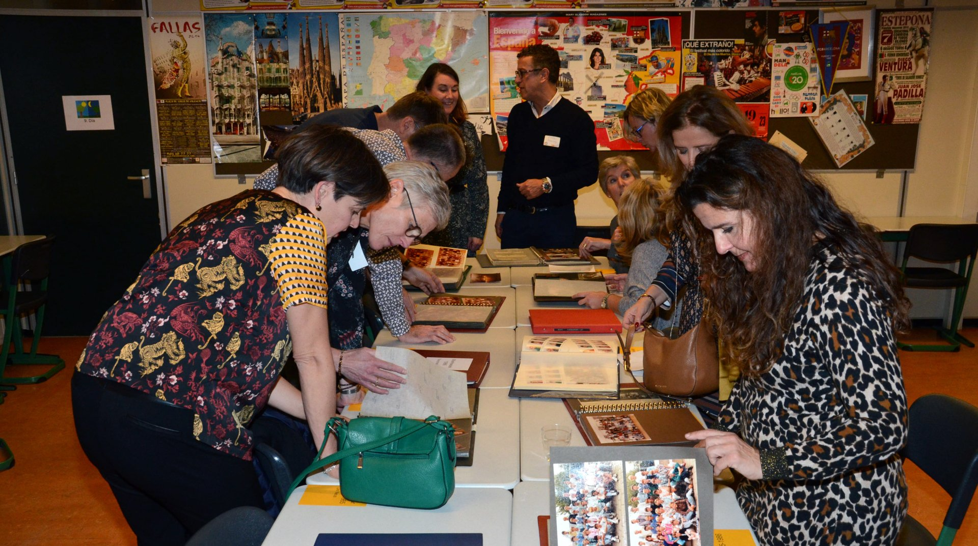 Mensen bekijken foto's op een tafel in een klaslokaal met posters aan de muur.