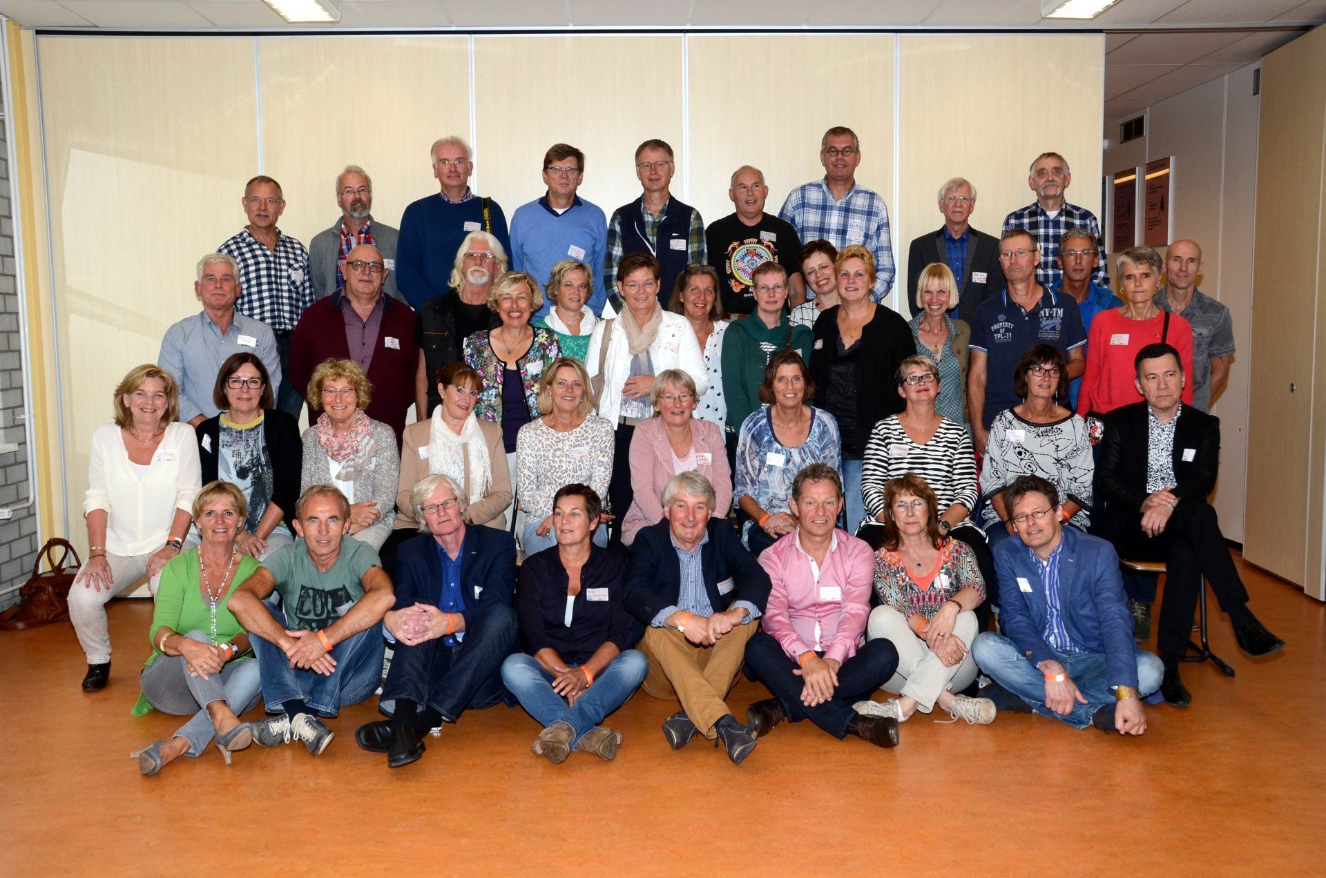Een groep mensen poseert voor een foto in een kamer. De meesten glimlachen, een paar zitten, neutrale achtergrond.