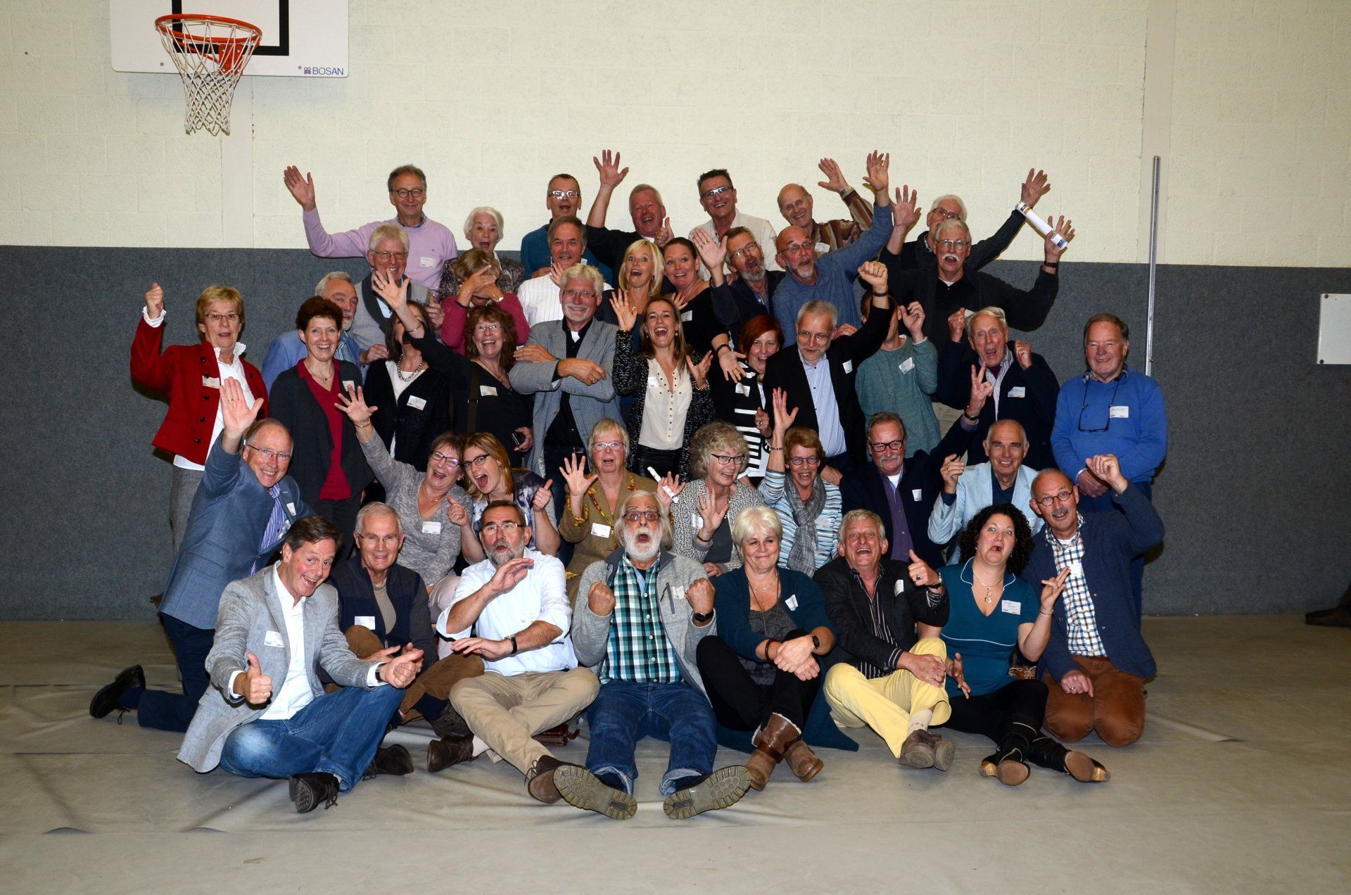 Een grote groep mensen in een gymzaal lacht en zwaait naar de camera.