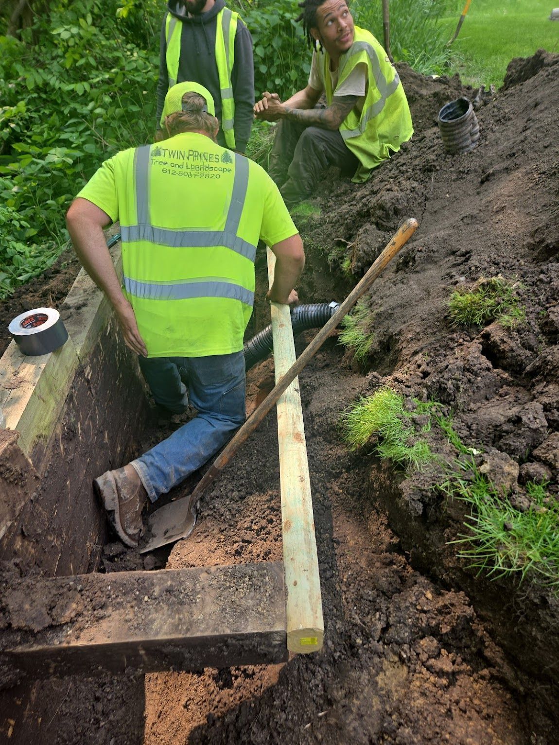 Three workers in high-visibility vests work on a construction site, installing wooden supports in a muddy trench.