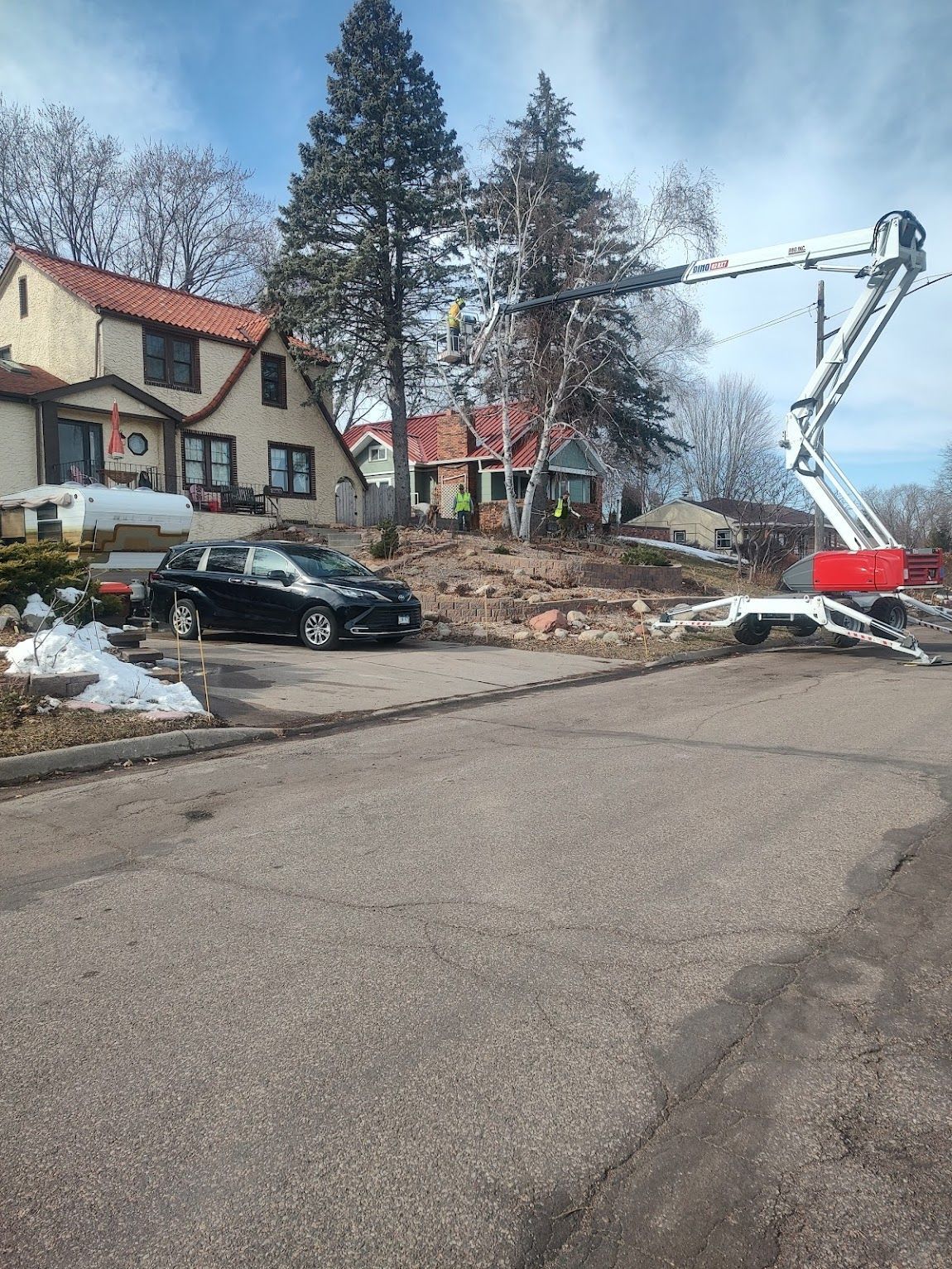 A utility truck with a raised boom arm works on trees in front of two houses on a sunny residential street.