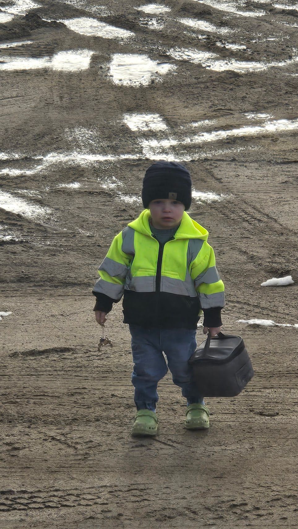 A child in a high-visibility yellow and black jacket and a black beanie walks across a muddy, rutted field holding a bag.