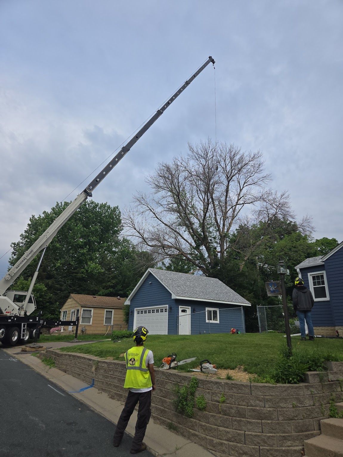A crane extends its boom over a suburban yard as workers prepare for tree removal near a blue garage.