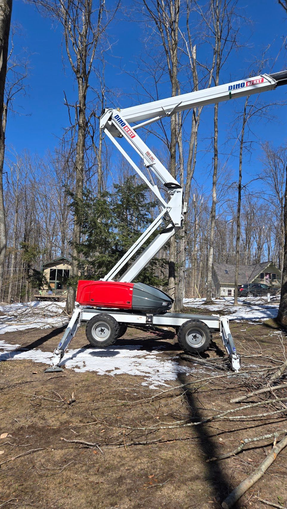 A white and red articulating boom lift standing in a wooded area with patches of snow on the ground under a clear blue sky.
