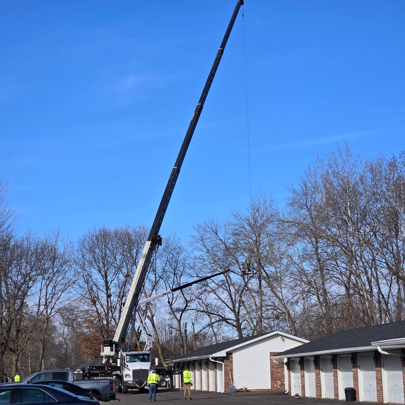 A large crane is parked in a parking lot, lifting a worker in a harness toward tall trees near a row of garage units.