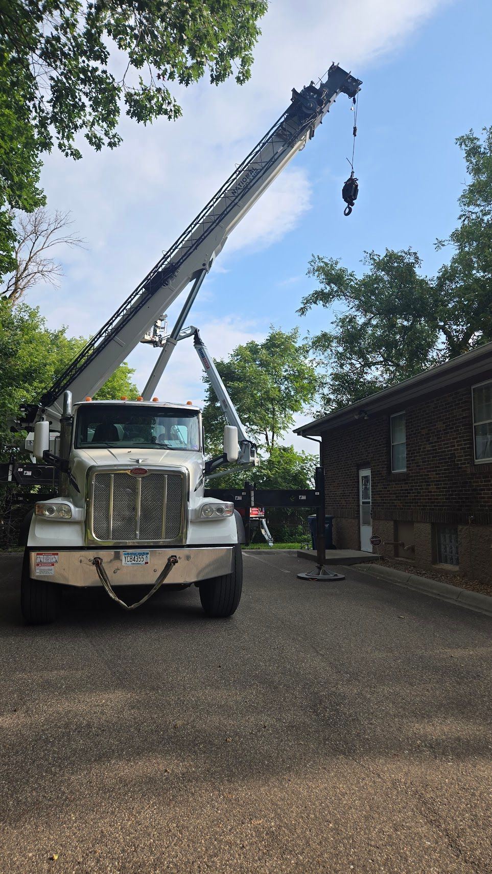 A large white crane truck is parked on a gravel driveway next to a brick house under a blue sky with trees.