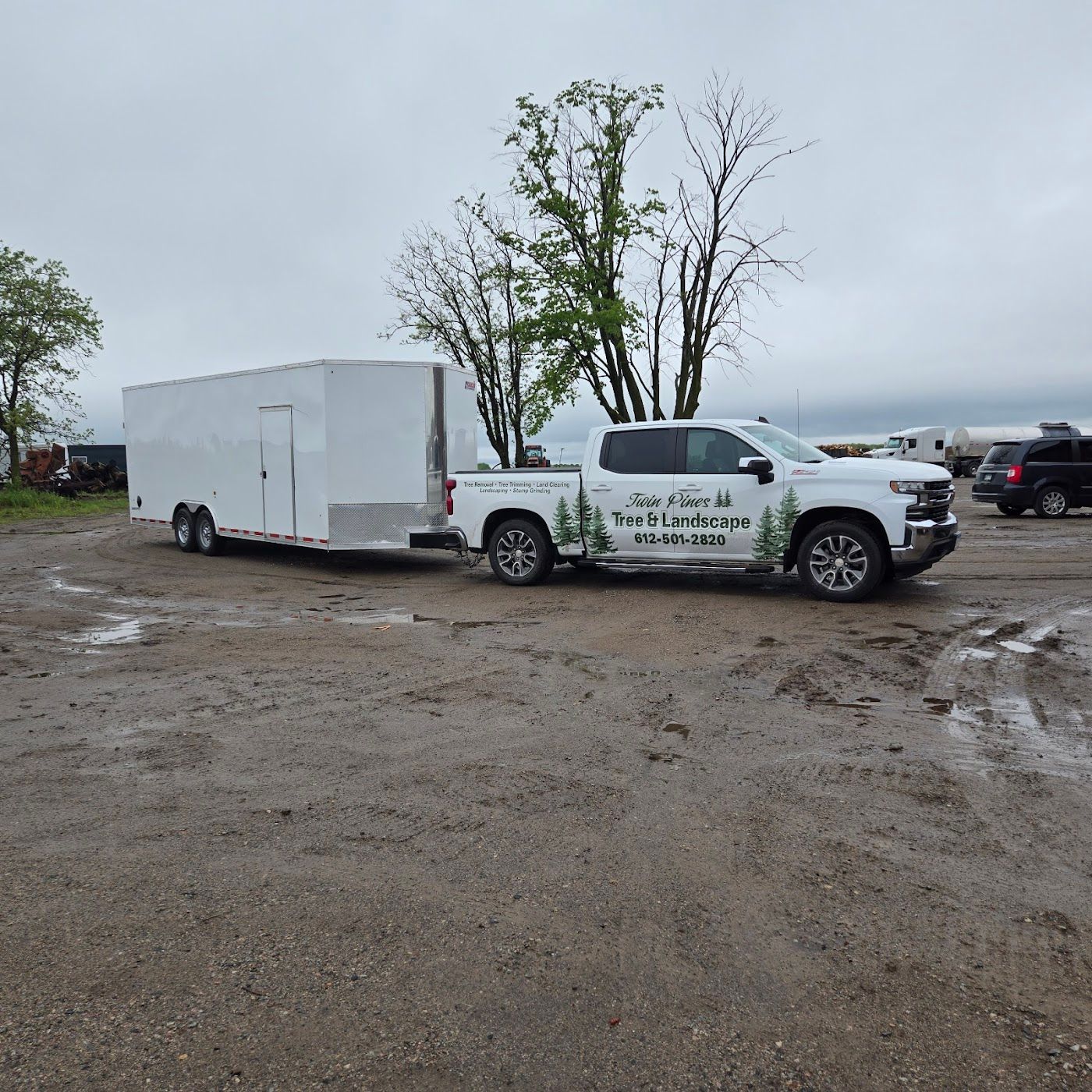 A white landscape service pickup truck towing a matching white enclosed trailer on a muddy lot under a cloudy sky.