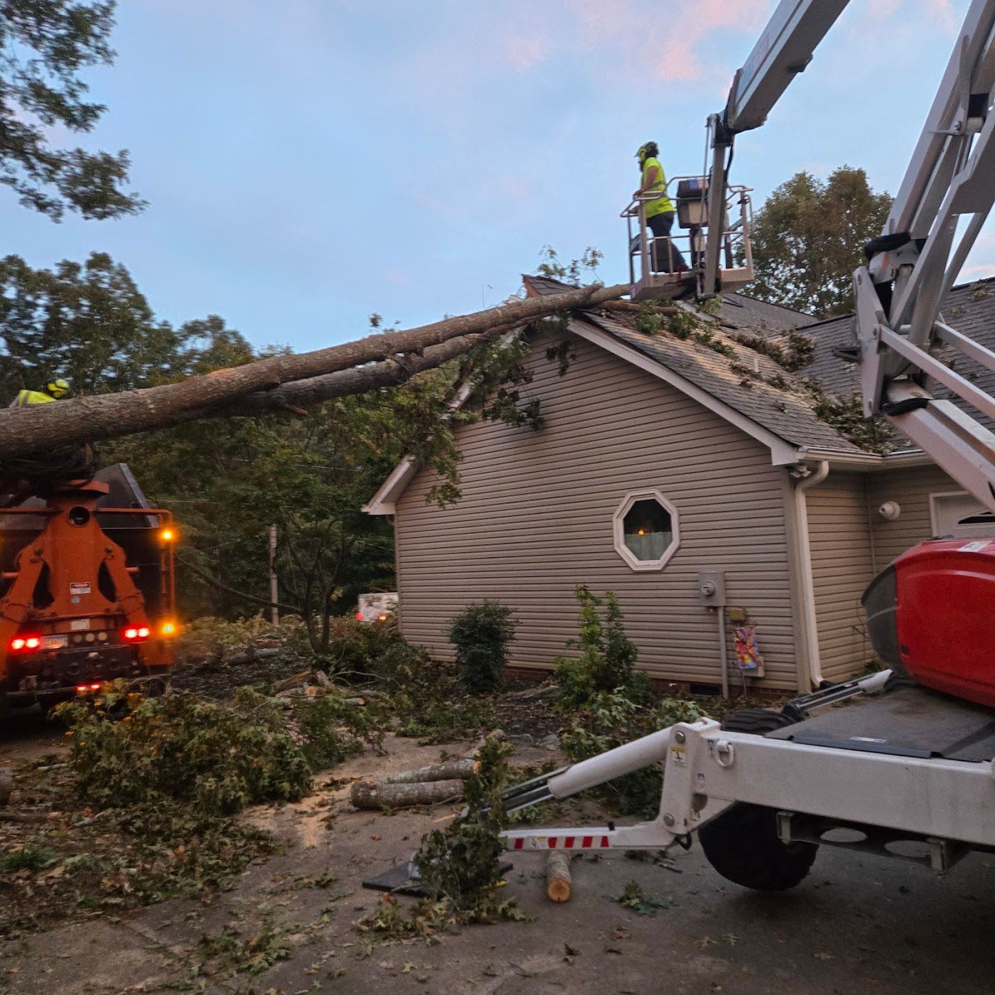 A tree removal crew uses a bucket truck to clear a large fallen tree from the roof of a beige house.