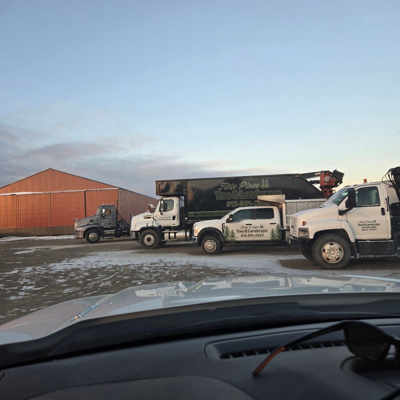Three trucks are parked in a snow-dusted lot beside a large red metal building under a clear blue sky.