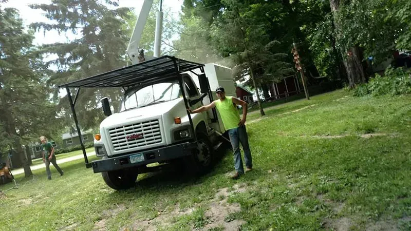 A person in a neon yellow shirt stands next to a white GMC bucket truck in a grassy, tree-filled area.