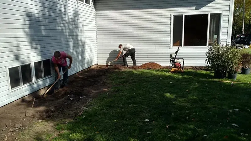 Two individuals work on a trench along the exterior foundation of a white-sided house on a sunny day.