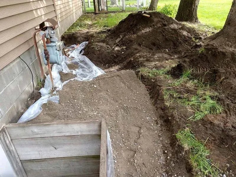 Excavated trench next to a home foundation with a gas meter, loose soil, and protective plastic sheeting.
